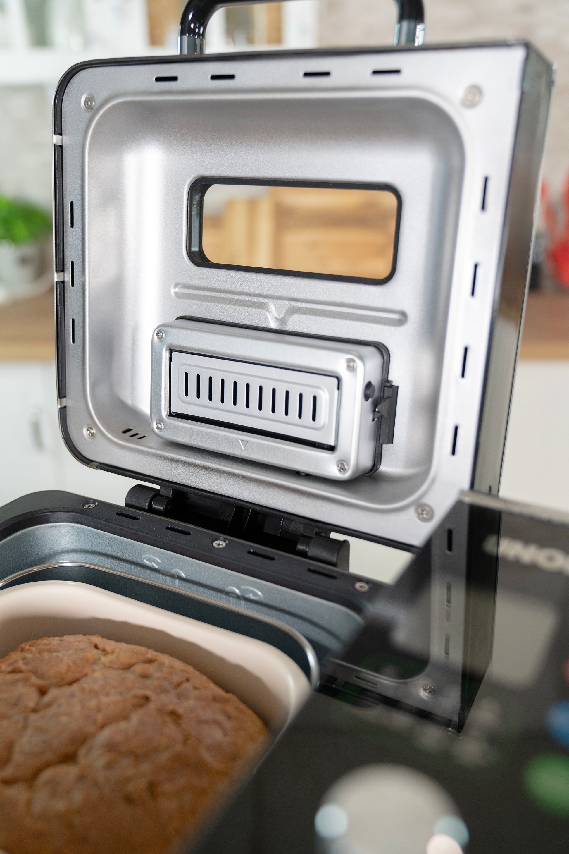 Der Unold AG Brotbackautomat BackProfi Nussspender mit geöffnetem Metalldeckel und Lüftungsschlitz offenbart ein frisch gebackenes Brot im Inneren. Der unscharfe Küchenhintergrund hebt das mitgelieferte Rezeptheft der Unold AG hervor.