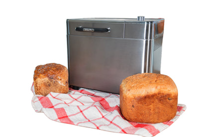 Der Unold AG Brotbackautomat BackProfi Nussspender inkl.. Rezeptheft sitzt auf einem rot-weiß-karierten Tuch, davor zwei Brote. Der Hintergrund ist einfarbig weiß.