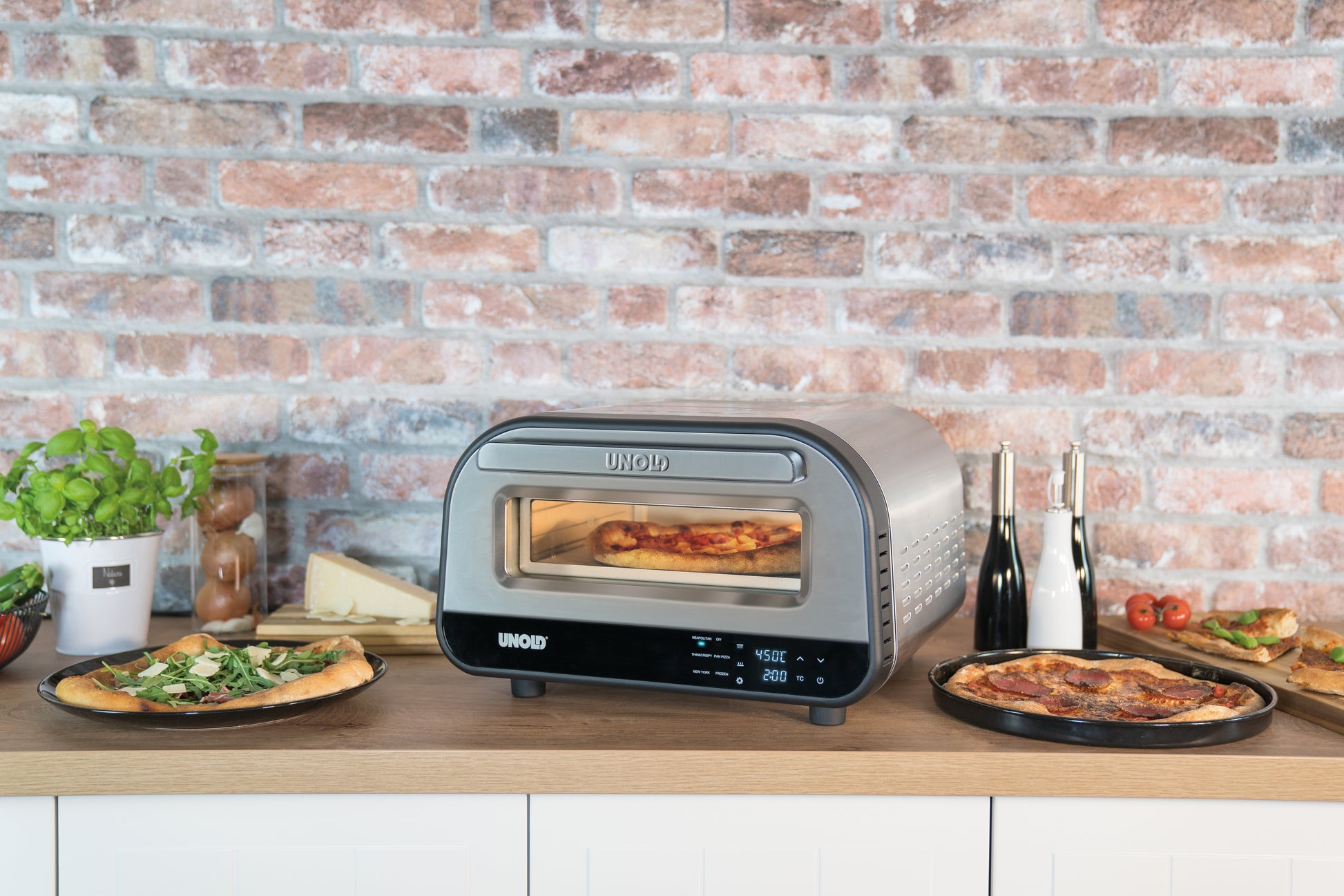 Der Unold Pizzaofen LUIGI steht kompakt auf einer Küchentheke mit einer Backsteinwand im Hintergrund. Frisch gebackene Pizzen liegen neben dem Ofen, umgeben von Kräutern, Gewürzen und Gemüse für eine schmackhafte Note.