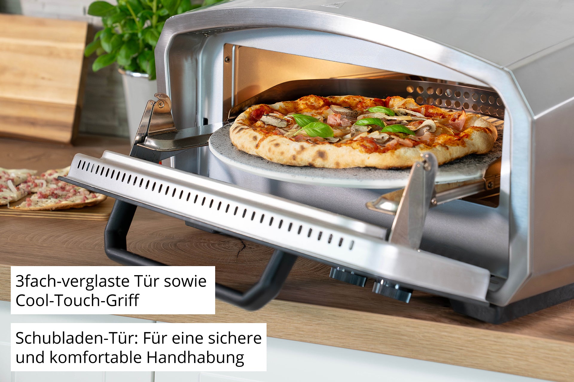 Der Unold Pizzaofen Enzo mit Metallgehäuse und Glastür steht offen auf einer hölzernen Arbeitsplatte, in der eine Pizza mit Gemüse und Basilikum zubereitet wird. Eine weitere Pizza ist im Hintergrund zu sehen.
