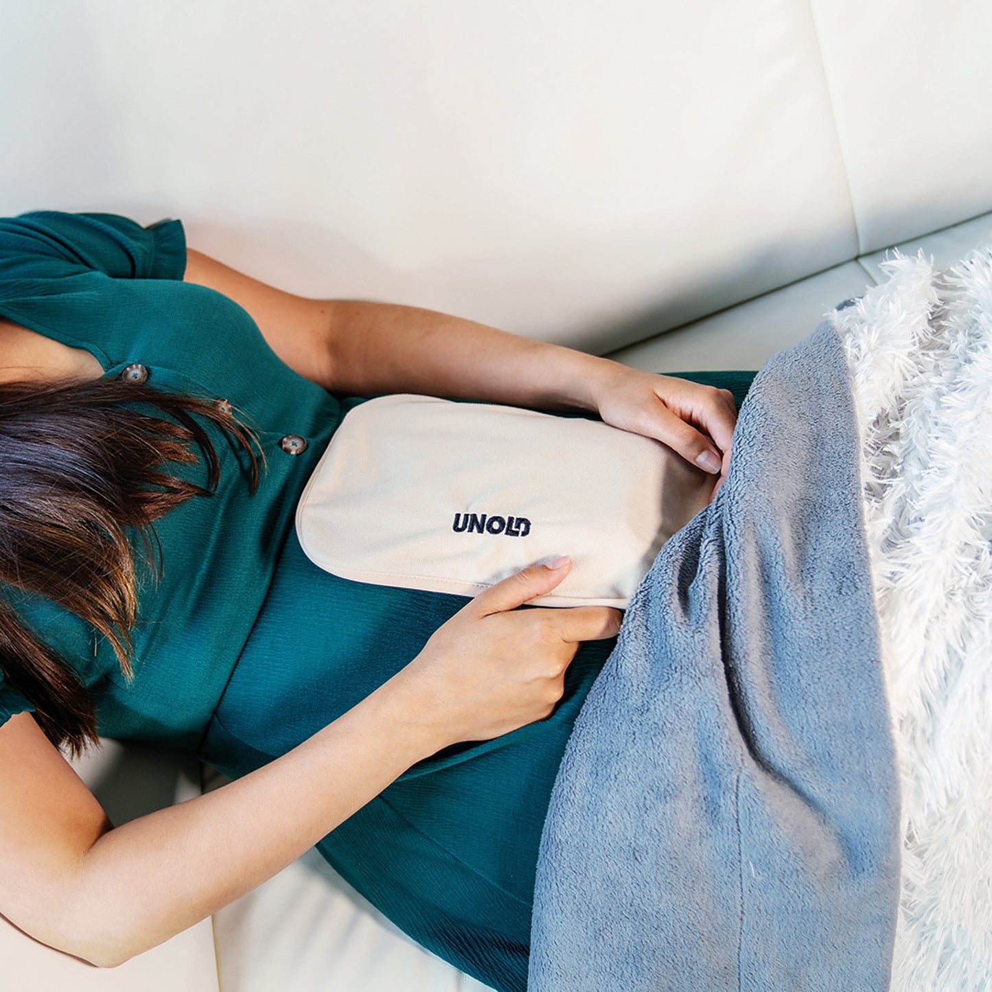Eine grün gekleidete Person liegt auf einem weißen Sofa und hält sich das Unold AG Wärmi Premium Set Heizkissen auf den Bauch, teilweise zugedeckt von einer grauen Decke.