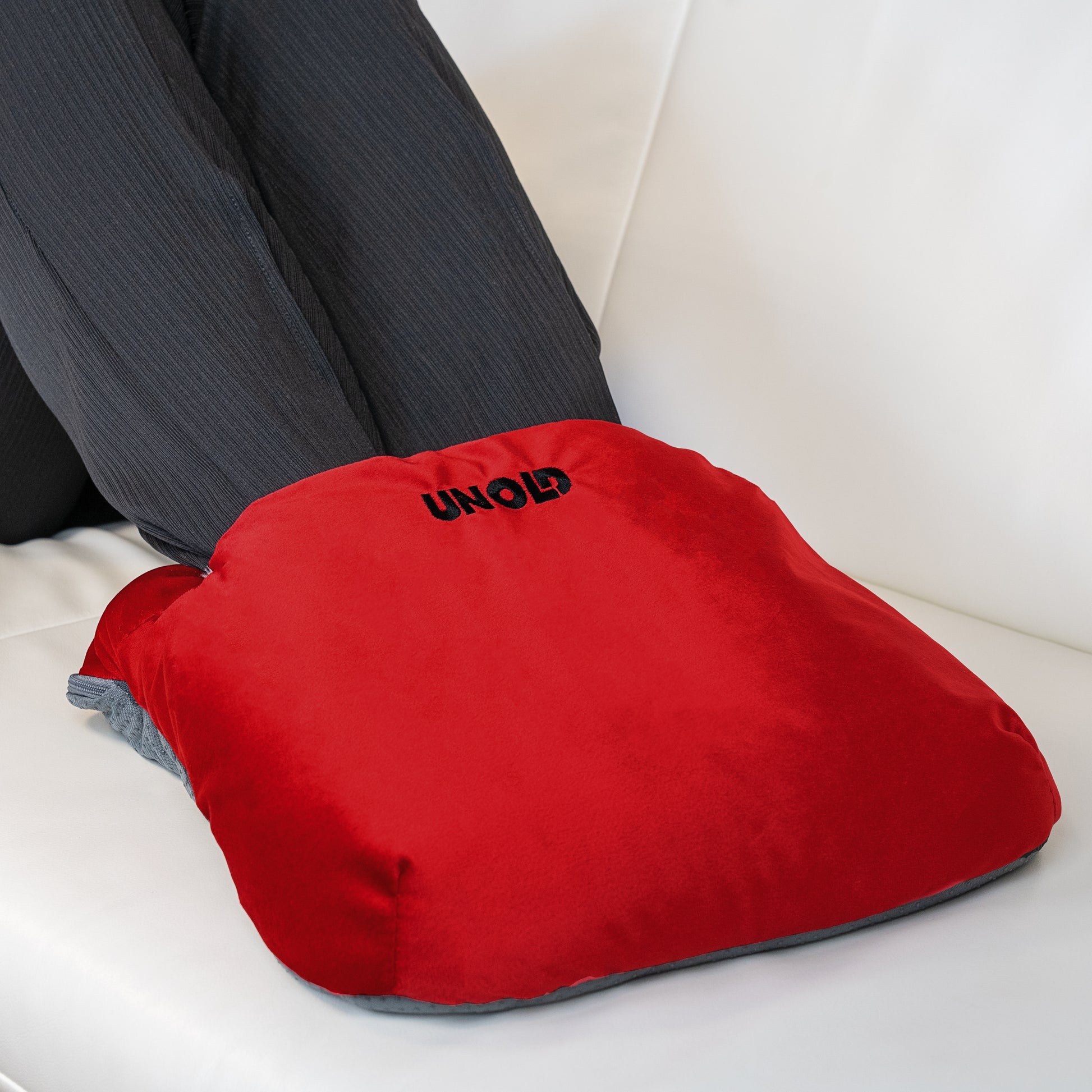 Eine Person in dunkler Hose entspannt sich auf einem weissen Sofa, ihre Füsse stecken in einem roten Wärmi Premium Set Fusswärmer der Unold AG, auf dem der Markenname prangt.
