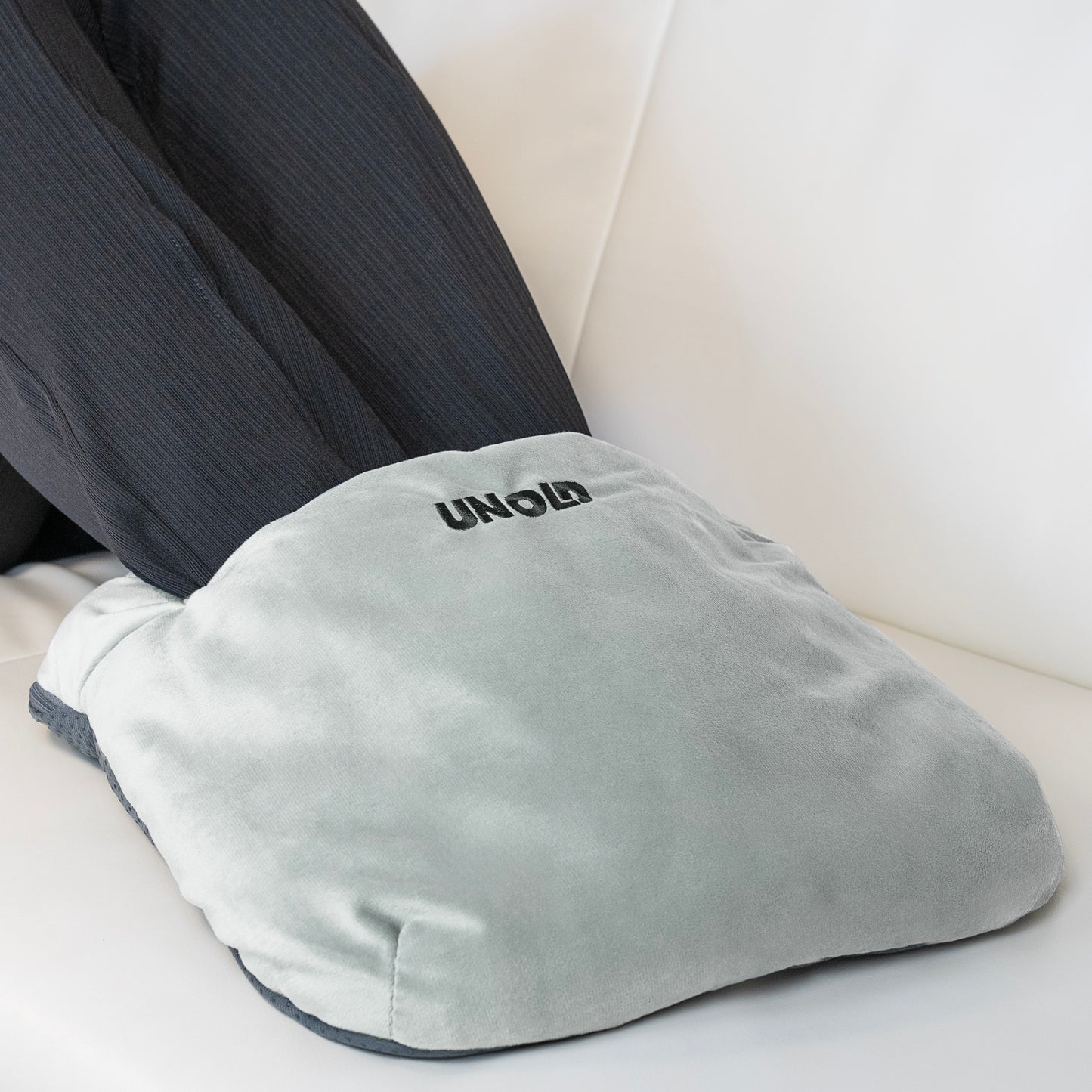 Eine Person, die eine dunkle Hose trägt, hat ihre Füße in einem weichen, grauen Unold 86030/86033/86037 Fusswärmekissen einzeln, das auf einer weißen Fläche ruht.