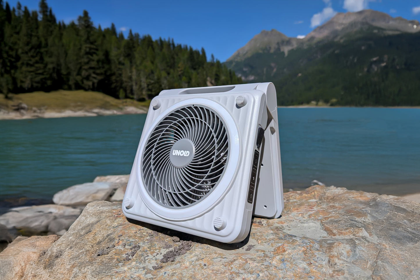 Ein kompakter, weißer Unold Solarventilator Camping-Ventilator ruht auf einem großen Felsen neben einem türkisfarbenen See, umgeben von Kiefern und Bergen unter einem klaren blauen Himmel.