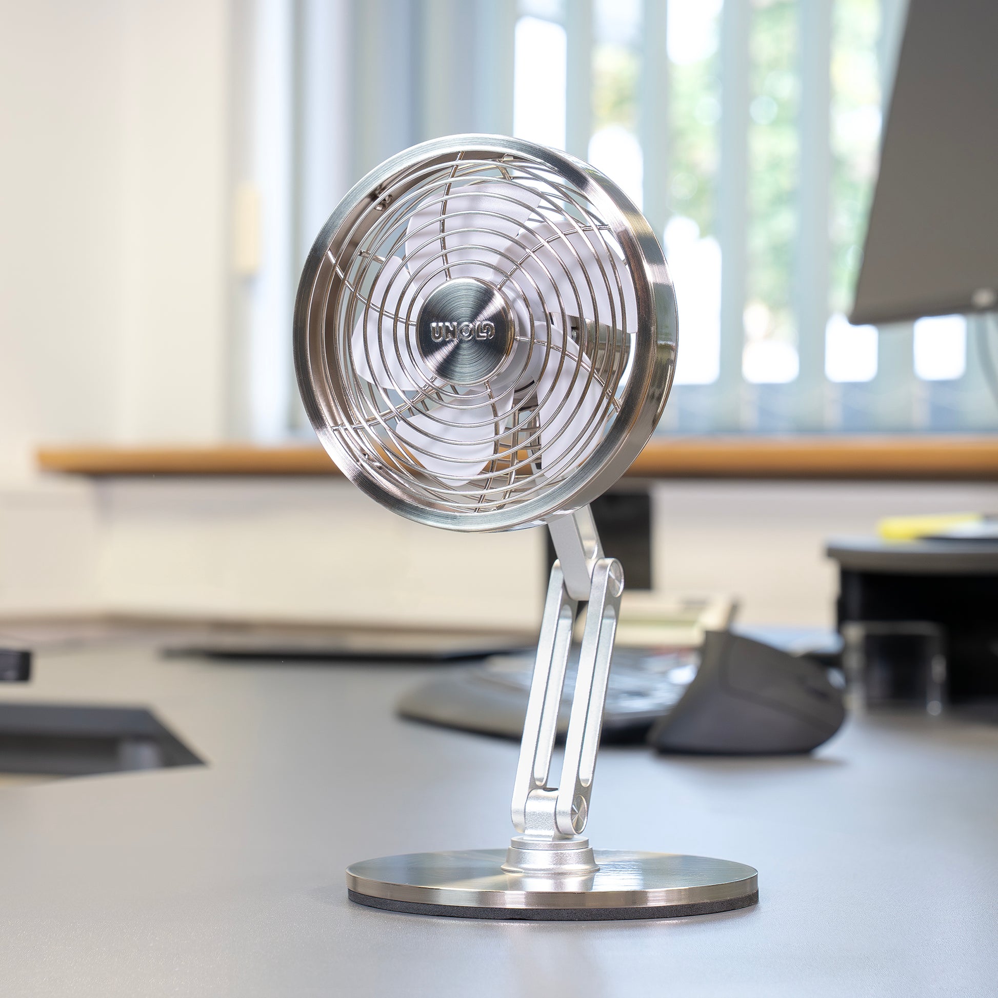 Der Unold AG Akku-Tischventilator Steel Flex, ein kompakter und moderner silberner Tischventilator, steht auf einem Bürotisch neben einer Computertastatur und einem Monitor in der Nähe eines sonnenbeschienenen Fensters mit Vertikaljalousien.