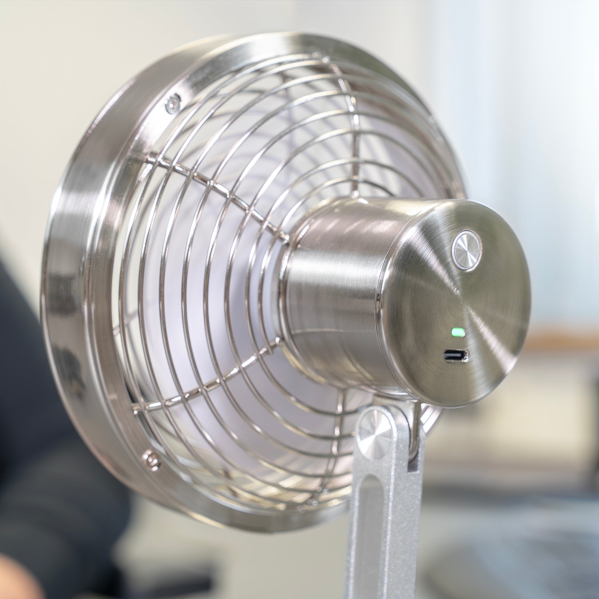 Eine Nahaufnahme des Unold AG Akku-Tischventilator Steel Flex, ein Tischventilator aus Edelstahl mit USB-Anschluss und Kontrollleuchte, platziert auf einem Bürotisch mit unscharfem Hintergrund.