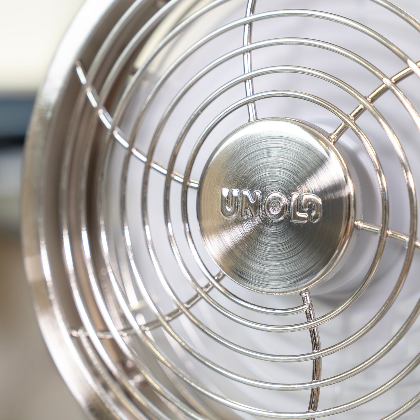 Die Nahaufnahme des Akku-Tischventilators Steel Flex der Unold AG zeigt das metallische Lüftergitter mit geprägtem Branding. Die glänzende Metalloberfläche und die runden Drähte unterstreichen das moderne, industrielle Design.