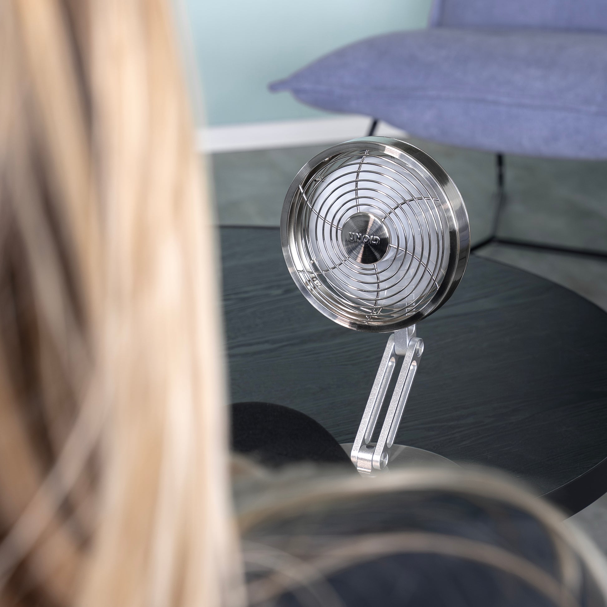Eine Person mit langen blonden Haaren sitzt vor dem Unold AG Akku-Tischventilator Steel Flex auf einem schwarzen Tisch, mit einem blauen Stuhl und einer hellblauen Wand im Hintergrund.