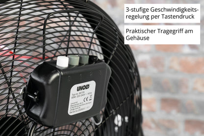 Nahaufnahme des schwarzen Schaltkastens der Unold Windmaschine Black Steel auf einem Gebläsegitter, mit drei beschrifteten Knöpfen für die 3-Stufen-Regelung und einem Tragegriff. Ein deutscher Text erklärt die Funktionen; im Hintergrund ist eine unscharfe Backsteinmauer zu sehen.