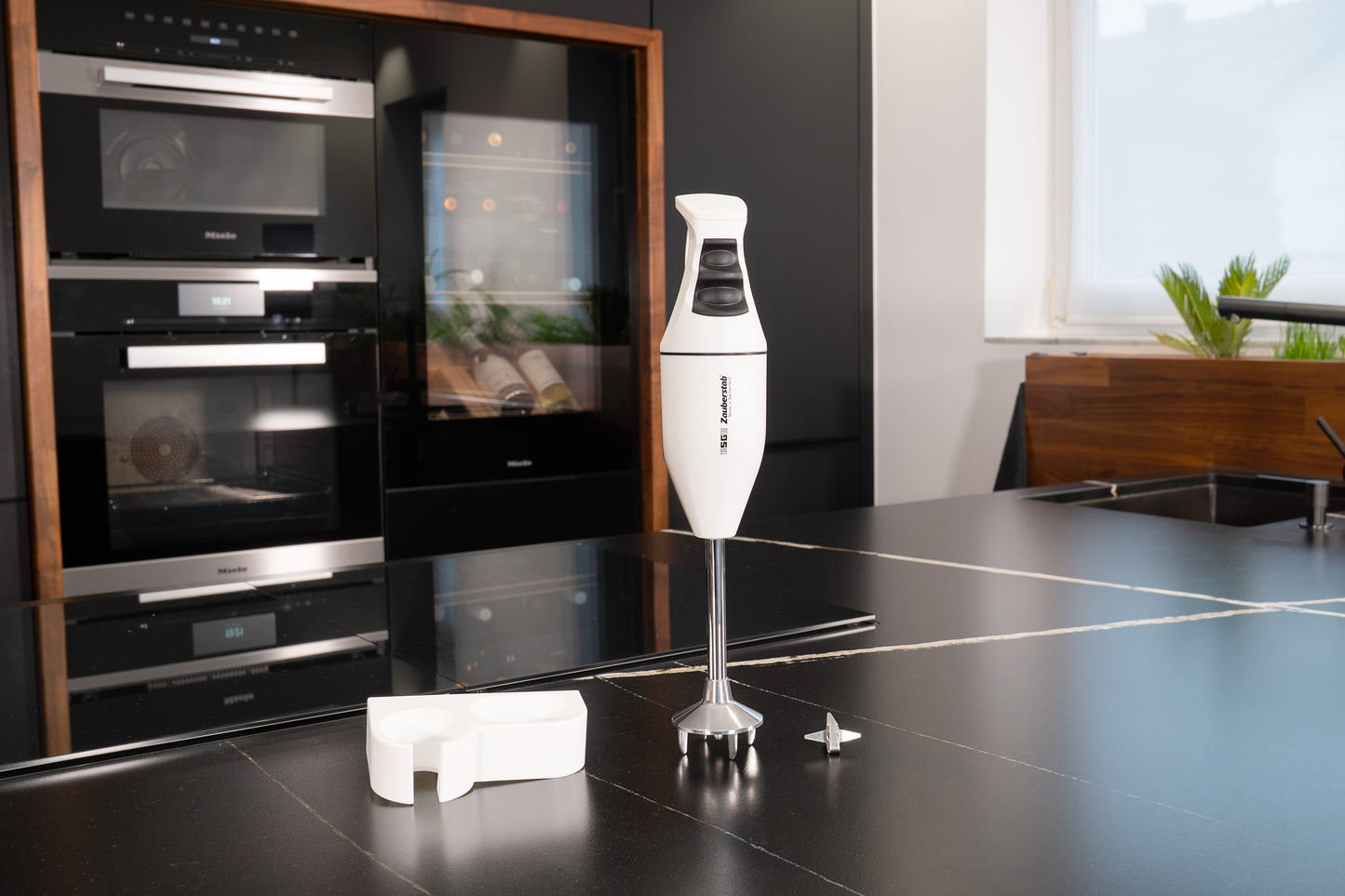 Der Unold ESGE-Zauberstab® M 100 Design Weiß Stabmixer mit Ständer und Metallklinge steht auf einer modernen schwarzen Küchenarbeitsplatte, auf der im Hintergrund ein Backofen und ein Weinkühlschrank zu sehen sind.