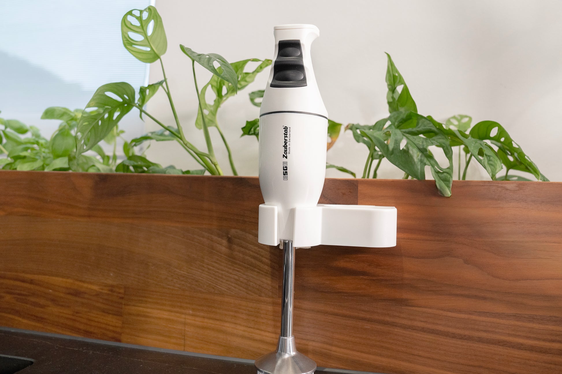Der Unold ESGE-Zauberstab® M 100 Design Weiß ist an einer hölzernen Küchenwand montiert, mit grünen Blattpflanzen im Hintergrund.