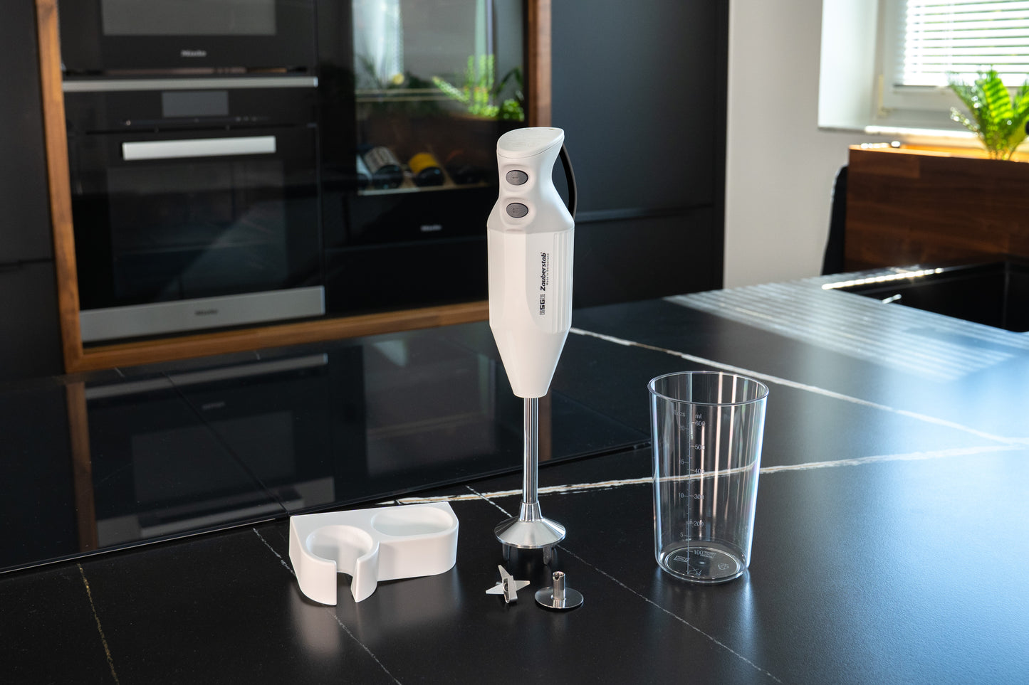 Der ESGE-Zauberstab® M 122 S von ESGE-Zauberstab, ein weißer Stabmixer mit Aufsätzen - Messbecher, Klinge und Aufbewahrungsbehälter - steht auf einer schwarzen Küchenarbeitsplatte in einer modernen Küche.