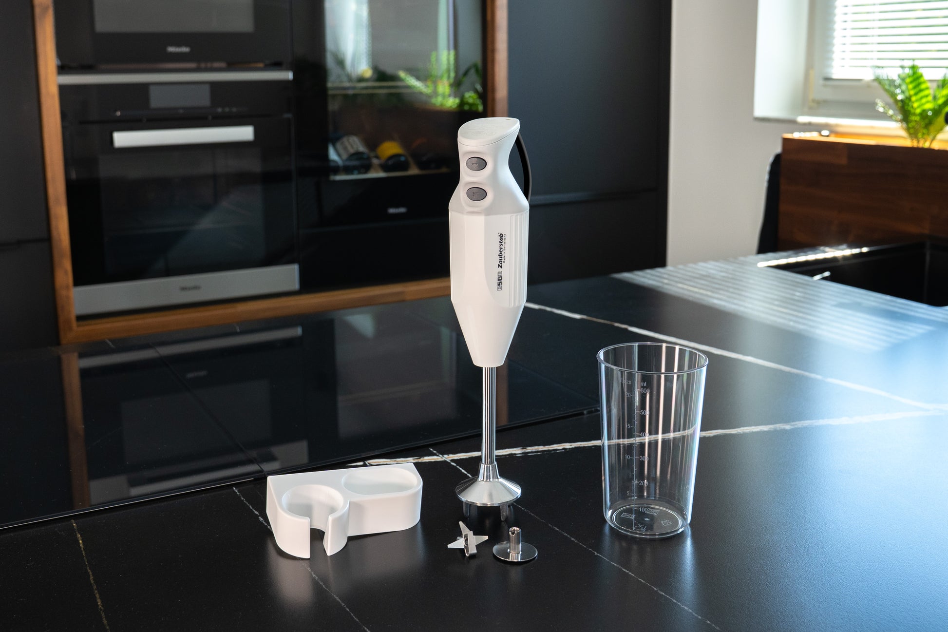 Der ESGE-Zauberstab® M 122 S von ESGE-Zauberstab, ein weißer Stabmixer mit Aufsätzen - Messbecher, Klinge und Aufbewahrungsbehälter - steht auf einer schwarzen Küchenarbeitsplatte in einer modernen Küche.
