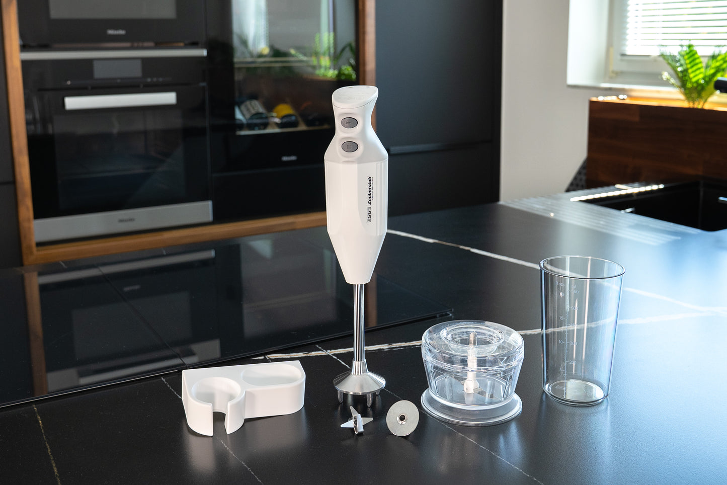 Der Unold ESGE-Zauberstab® M 122 De Luxe, ein weißer Stabmixer mit einem Becher, einem Schneebesen, einer Schüssel zum Zerkleinern und einem Messeraufsatz, wird auf einer modernen schwarzen Küchenzeile gezeigt, die vom Sonnenlicht aus einem nahe gelegenen Fenster beleuchtet wird.