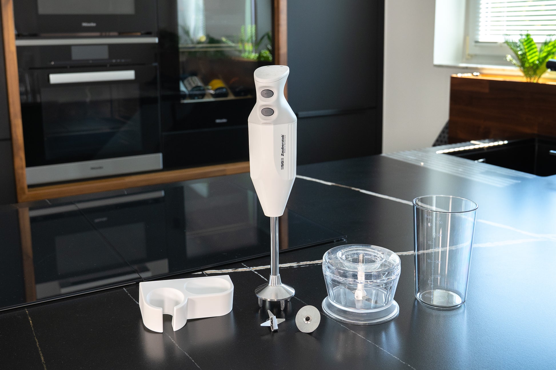 Der Unold ESGE-Zauberstab® M 122 De Luxe, ein weißer Stabmixer mit einem Becher, einem Schneebesen, einer Schüssel zum Zerkleinern und einem Messeraufsatz, wird auf einer modernen schwarzen Küchenzeile gezeigt, die vom Sonnenlicht aus einem nahe gelegenen Fenster beleuchtet wird.