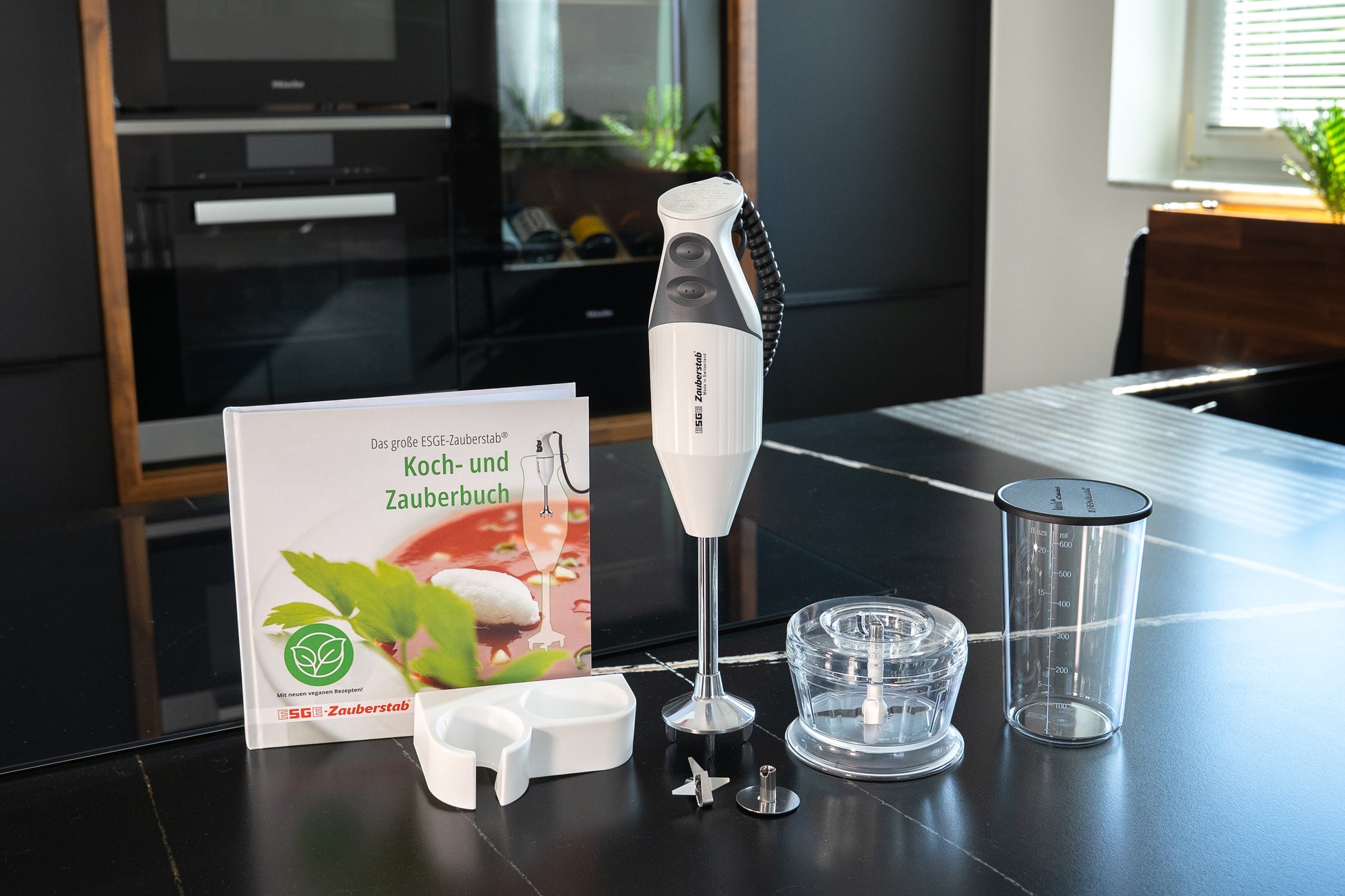 Der Unold ESGE-Zauberstab® M160 G Gourmet Stabmixer mit Aufsätzen, Messbecher, Zerkleinerungsschüssel, Wandhalterung und Kochbuch wird auf einer schwarzen Küchentheke mit einem Ofen und einem sonnendurchfluteten Fenster im Hintergrund gezeigt.