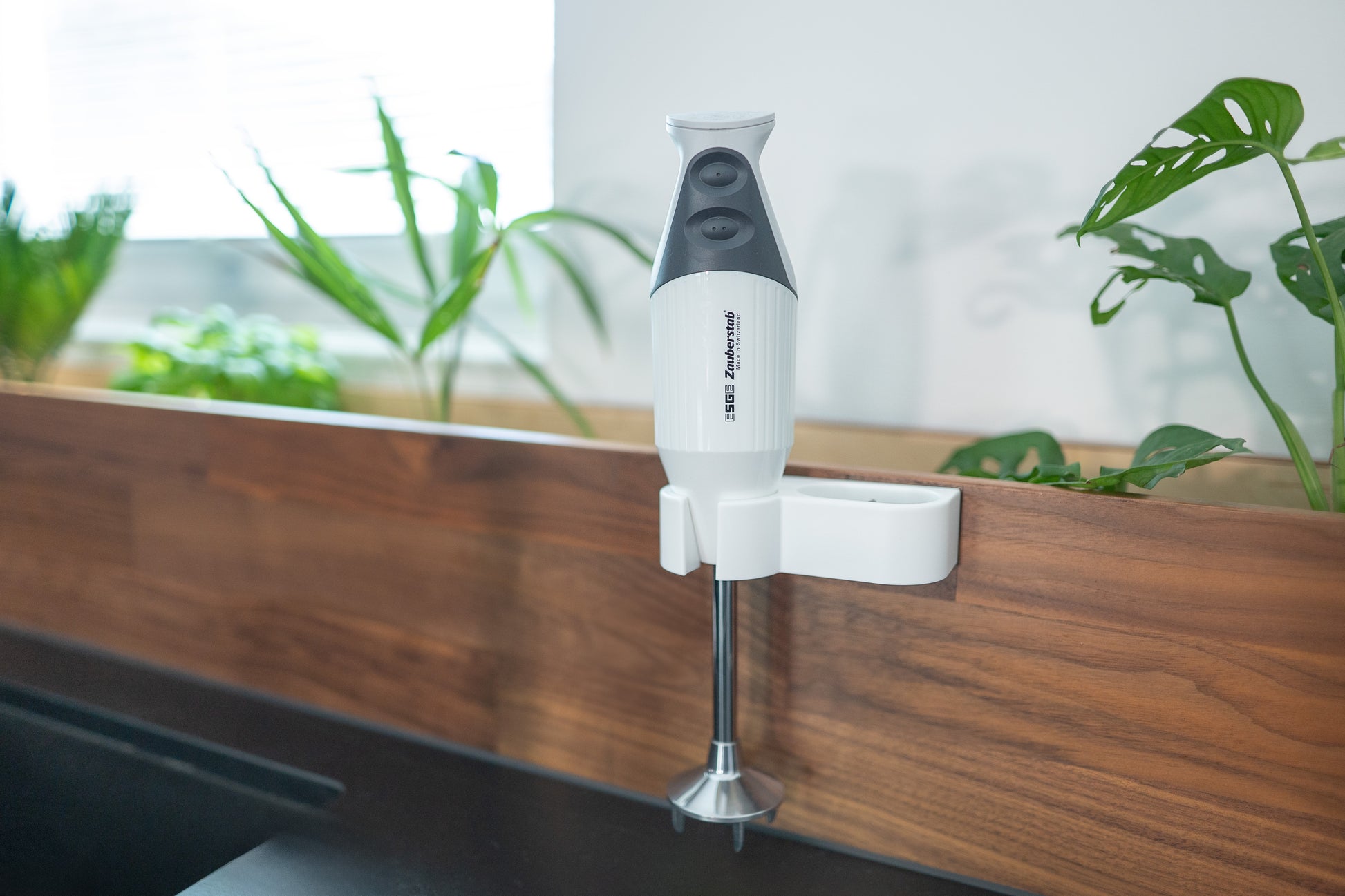 Der Unold ESGE-Zauberstab® M160 G Gourmet Stabmixer steht senkrecht auf einer hölzernen Küchenabdeckung, umgeben von grünen Topfpflanzen.