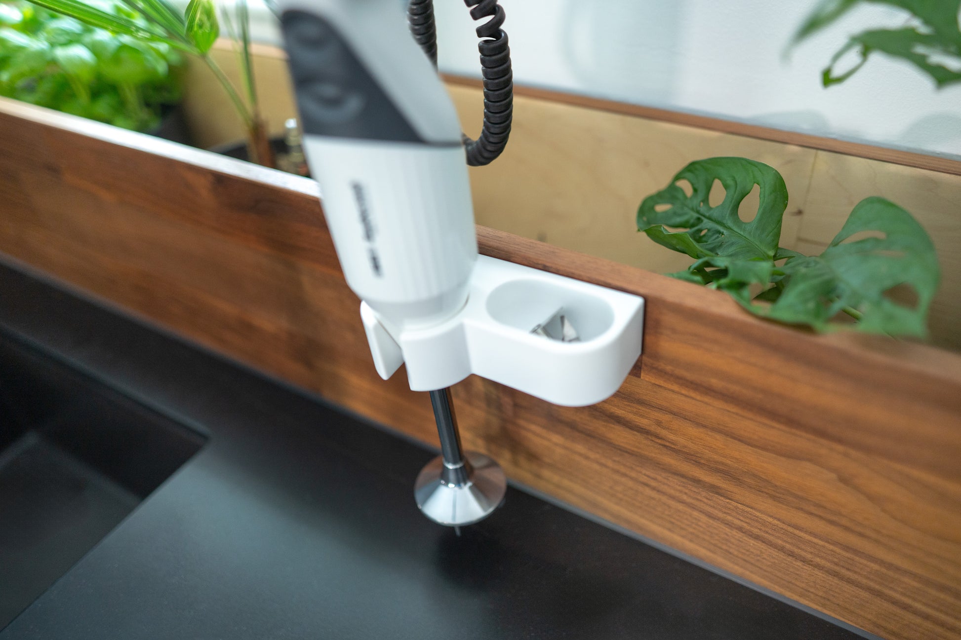 Ein ESGE-Zauberstab® M160 G Gourmet Stabmixer von Unold ist auf einer hölzernen Aufkantung über einer schwarzen Küchentheke montiert, mit Grünpflanzen im Hintergrund.