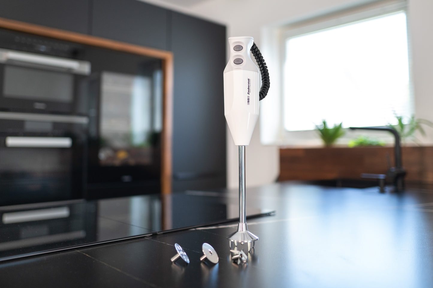 Der Unold ESGE-Zauberstab® P 200 Profi, ein weißer Stabmixer, steht aufrecht auf einem schwarzen Küchentisch mit seinen Metallaufsätzen in der Nähe in einer modernen Küche mit schwarzen Schränken und Einbaugeräten.