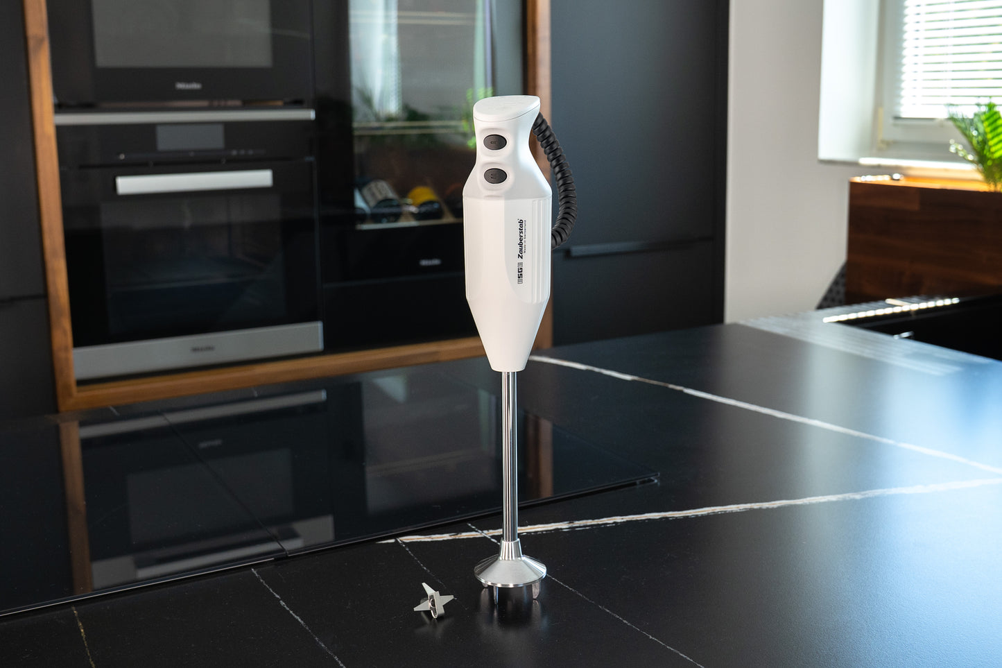 Der Unold ESGE-Zauberstab® P 350 Profi, ein weißer Stabmixer, steht aufrecht auf einer schwarzen Arbeitsplatte in einer modernen Küche, der kleine Metallklingenaufsatz liegt daneben.