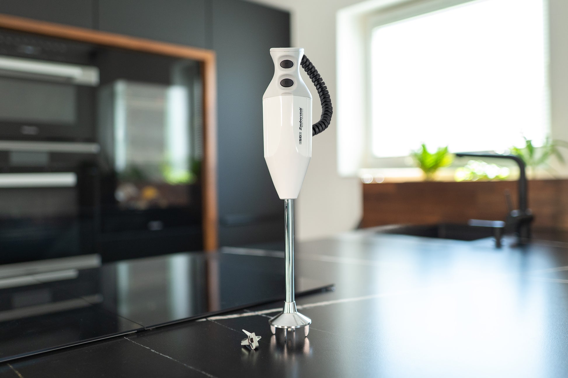 Der Unold ESGE-Zauberstab® P 350 Profi mit Metallschaft steht aufrecht auf einer schwarzen Arbeitsplatte in einer modernen Küche mit schwarzen Schränken, einem Fenster und Topfpflanzen im Hintergrund.