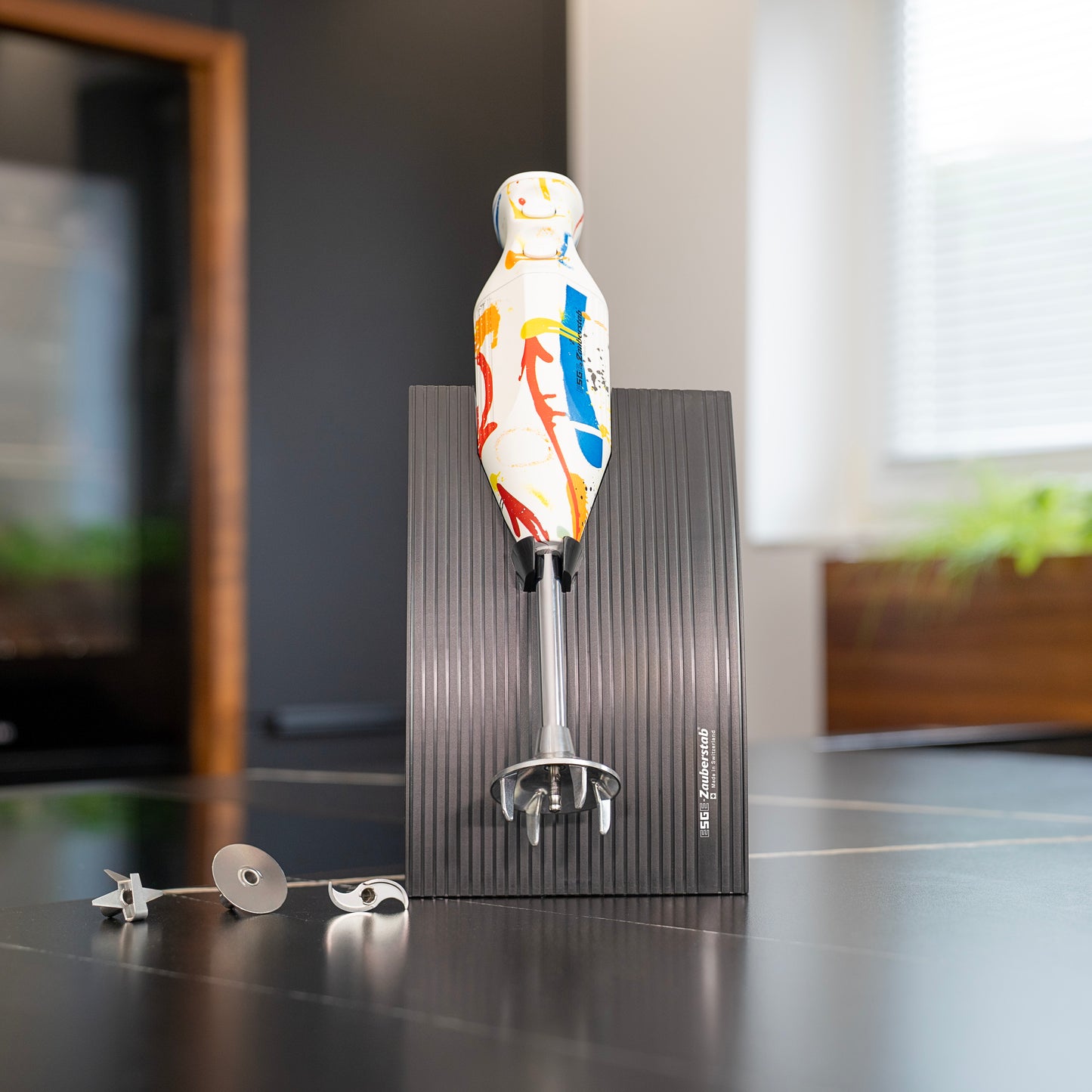 Der ESGE-Zauberstab Design Edition Color Art Stabmixer mit einem lebendigen, abstrakten Design steht aufrecht auf einem schwarzen, strukturierten Sockel mit seinen Aufsätzen in der Nähe auf einer Küchenzeile. Im Hintergrund sind ein Fenster und verschwommene Küchenelemente zu sehen.