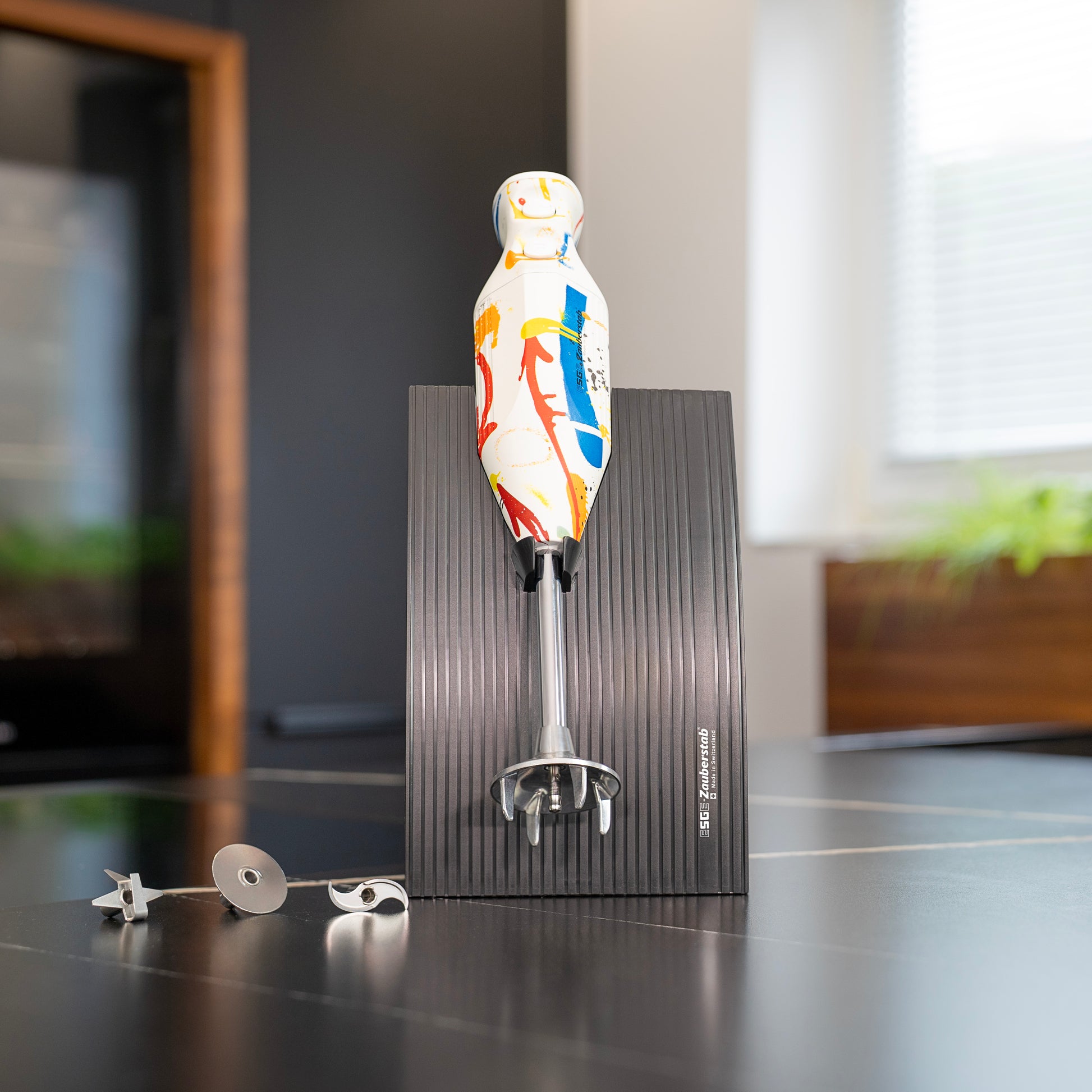 Der ESGE-Zauberstab Design Edition Color Art Stabmixer mit einem lebendigen, abstrakten Design steht aufrecht auf einem schwarzen, strukturierten Sockel mit seinen Aufsätzen in der Nähe auf einer Küchenzeile. Im Hintergrund sind ein Fenster und verschwommene Küchenelemente zu sehen.