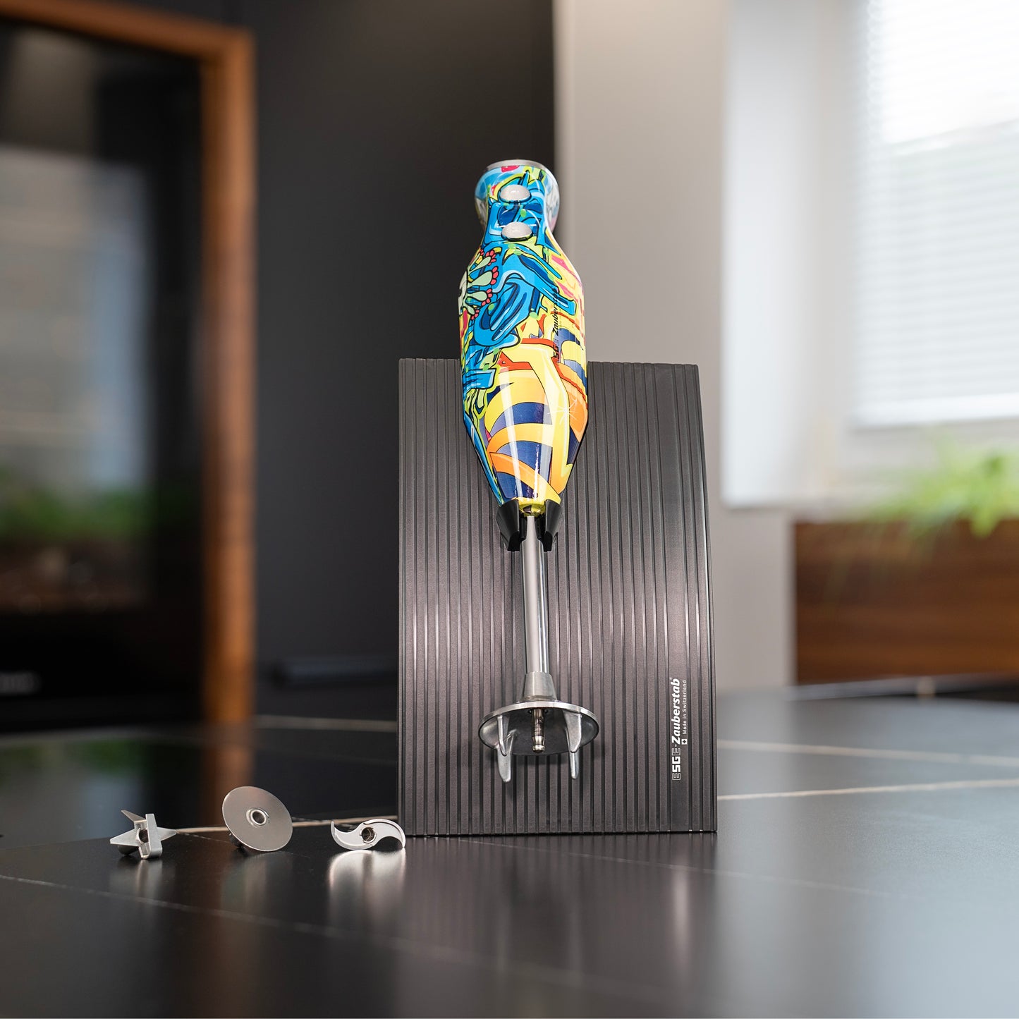 Der ESGE-Zauberstab Design Edition Pop Art Stabmixer von ESGE-Zauberstab mit Aufsätzen aus Edelstahl und schwarzem Ständer wird in einer lebendigen, modernen Küche mit verschwommenen Fenstern und Grünpflanzen im Hintergrund präsentiert.