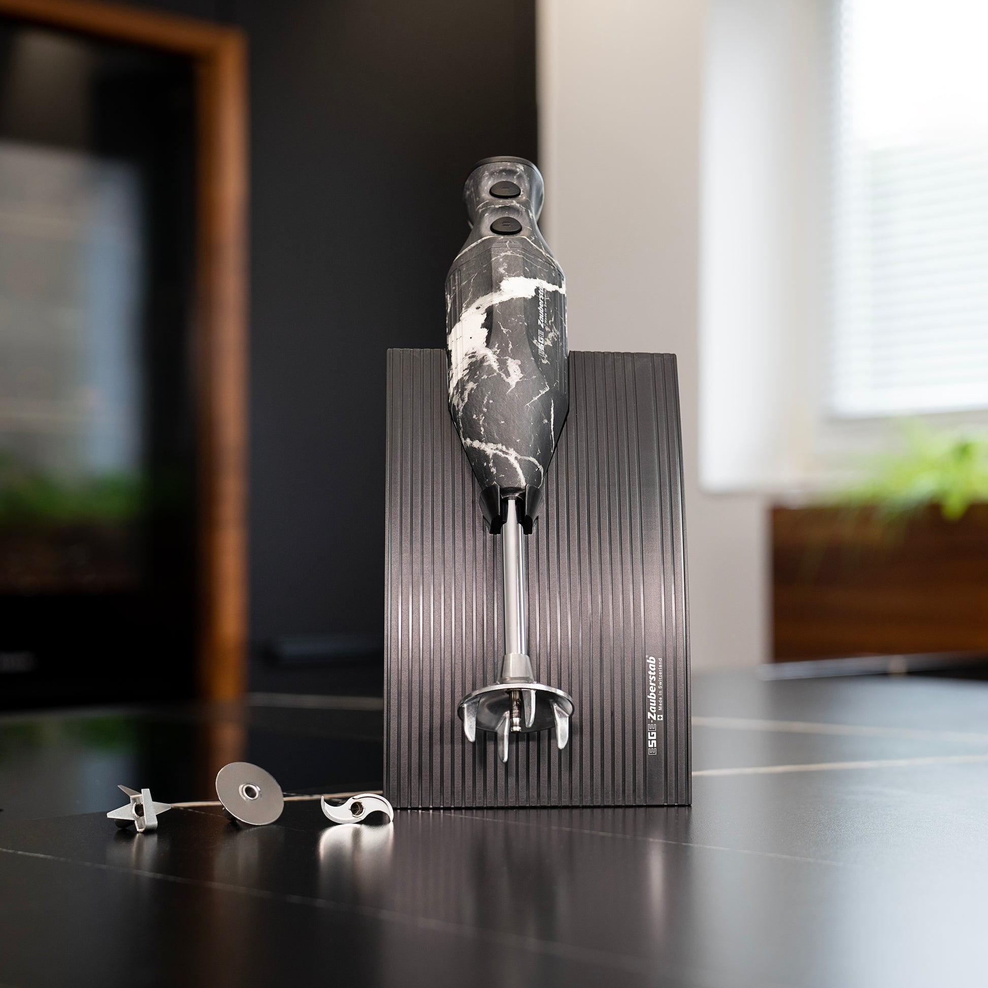 Der ESGE-Zauberstab Design Edition Marmor-Schwarz Stabmixer von ESGE-Zauberstab steht aufrecht auf einem gerippten Sockel, daneben drei Metallklingenaufsätze auf einer dunklen Küchenarbeitsplatte.
