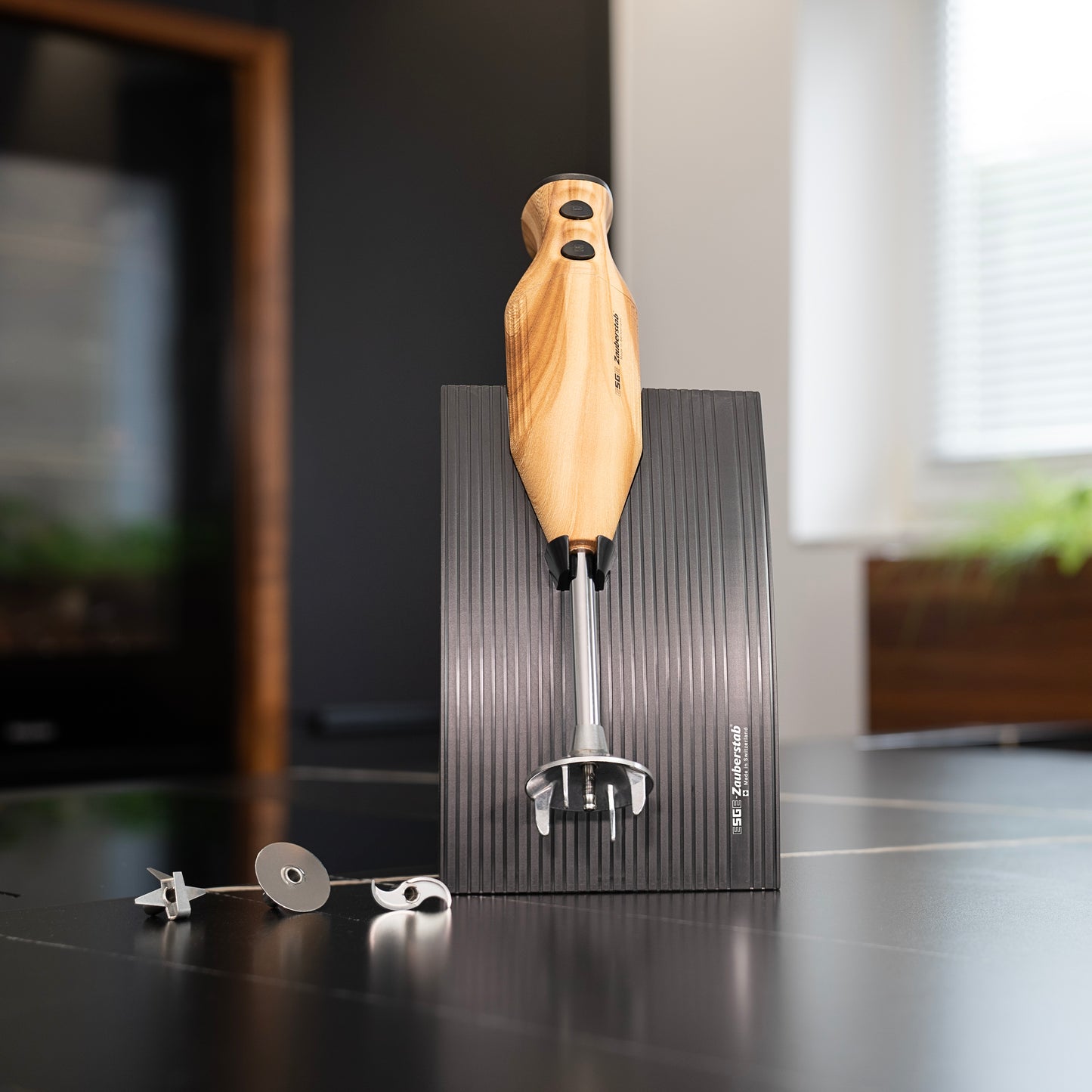 Der ESGE-Zauberstab Design Edition Douglasie Stabmixer von ESGE-Zauberstab steht aufrecht auf einem schwarzen gerillten Sockel mit drei Metallaufsätzen daneben auf einer modernen Küchentheke, mit einem Fenster und Pflanzen im Hintergrund.