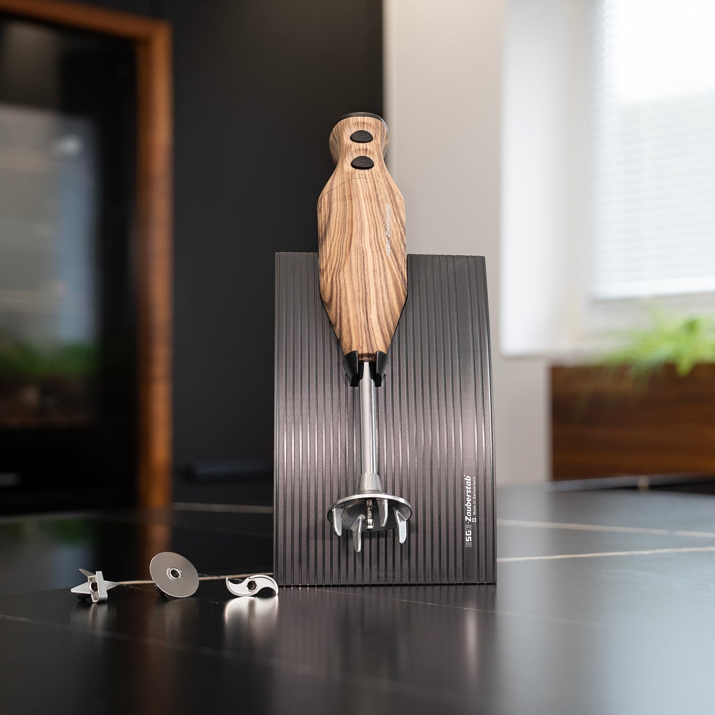 Der Stabmixer ESGE-Zauberstab Design Edition Nussbaum von ESGE-Zauberstab steht aufrecht auf einem schwarzen, gerippten Ständer, daneben mehrere Mixaufsätze aus Metall auf einer dunklen Arbeitsplatte in einer modernen Küche.