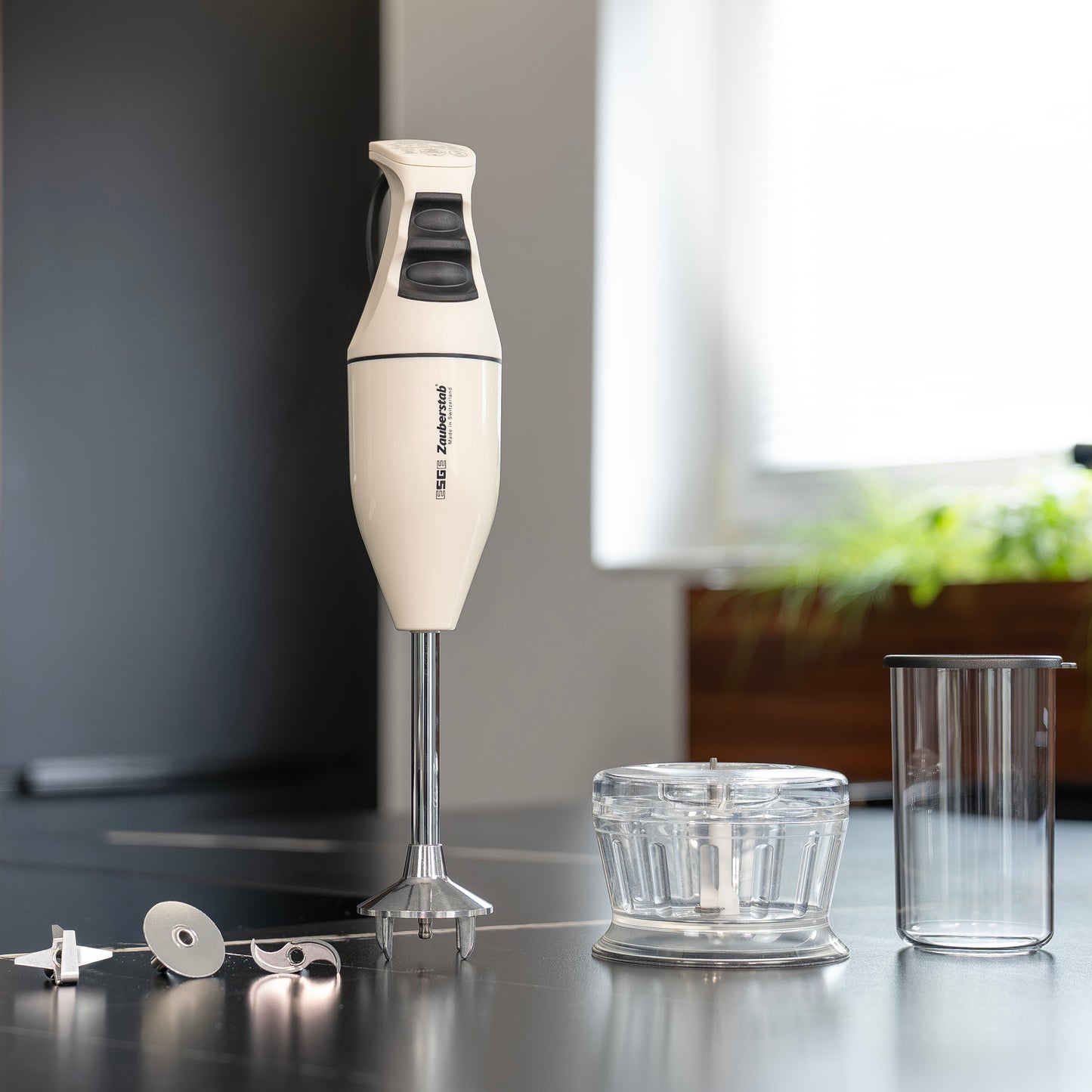 Der Unold ESGE-Zauberstab Bundle E 120 Select Creme, ein weißer Stabmixer mit schwarzen Akzenten, wird auf einem Küchentisch mit Aufsätzen, einer Schüssel zum Zerkleinern und einem durchsichtigen Messbecher gezeigt; im Hintergrund stehen Grünpflanzen.
