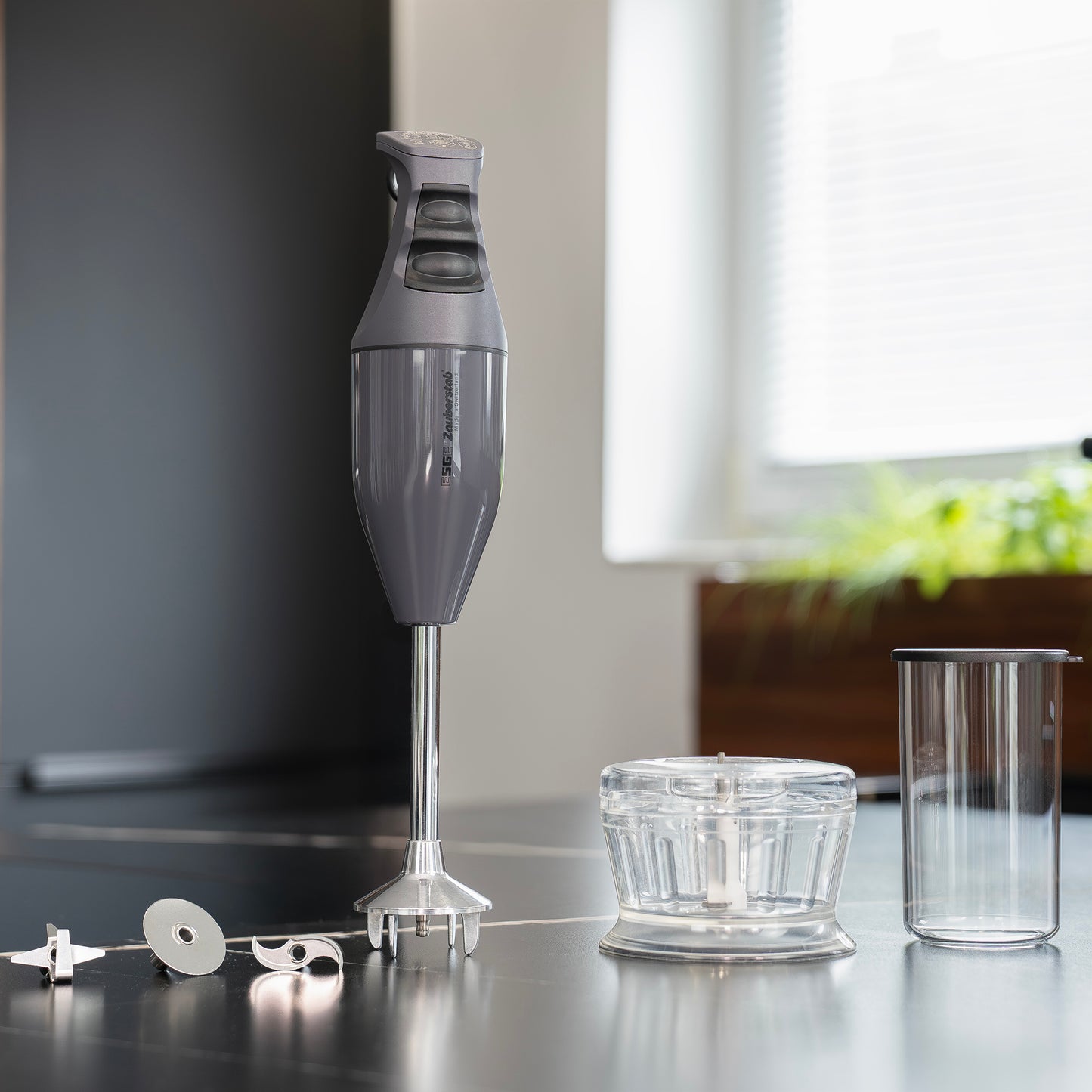 Der Unold ESGE-Zauberstab Bundle E 120 Select Anthrazit Stabmixer mit Aufsätzen, transparenter Zerkleinerungsschale und Messbecher steht ordentlich auf einem Küchentisch mit Sonnenlicht und Grünpflanzen im Hintergrund.
