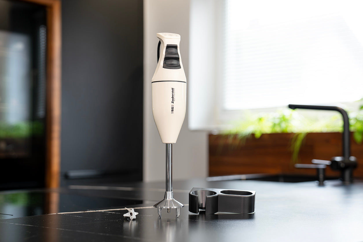 Der ESGE-Zauberstab M100 - Exklusiv Edition Creme Stabmixer von ESGE-Zauberstab steht aufrecht auf einer Küchentheke mit den Aufsätzen und der Halterung in der Nähe, während das Sonnenlicht aus einem Fenster grüne Pflanzen im Hintergrund hervorhebt.