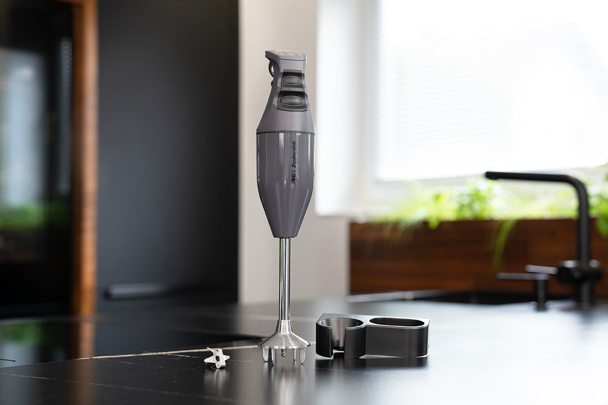 Der ESGE-Zauberstab M100 - Exklusiv Edition Anthrazit Stabmixer von ESGE-Zauberstab steht aufrecht mit seinen Aufsätzen auf einer schwarzen Küchentheke, während im Hintergrund das Sonnenlicht in eine moderne Küche fällt.