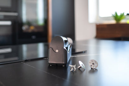 Eine Nahaufnahme des kabellosen Unold ESGE-Zauberstab® Schwarz 95105 auf einer Arbeitsplatte, mit drei Metallaufsätzen davor. Ein unscharfer Hintergrund zeigt einen Küchenherd und ein helles Fenster mit einer kleinen Topfpflanze.