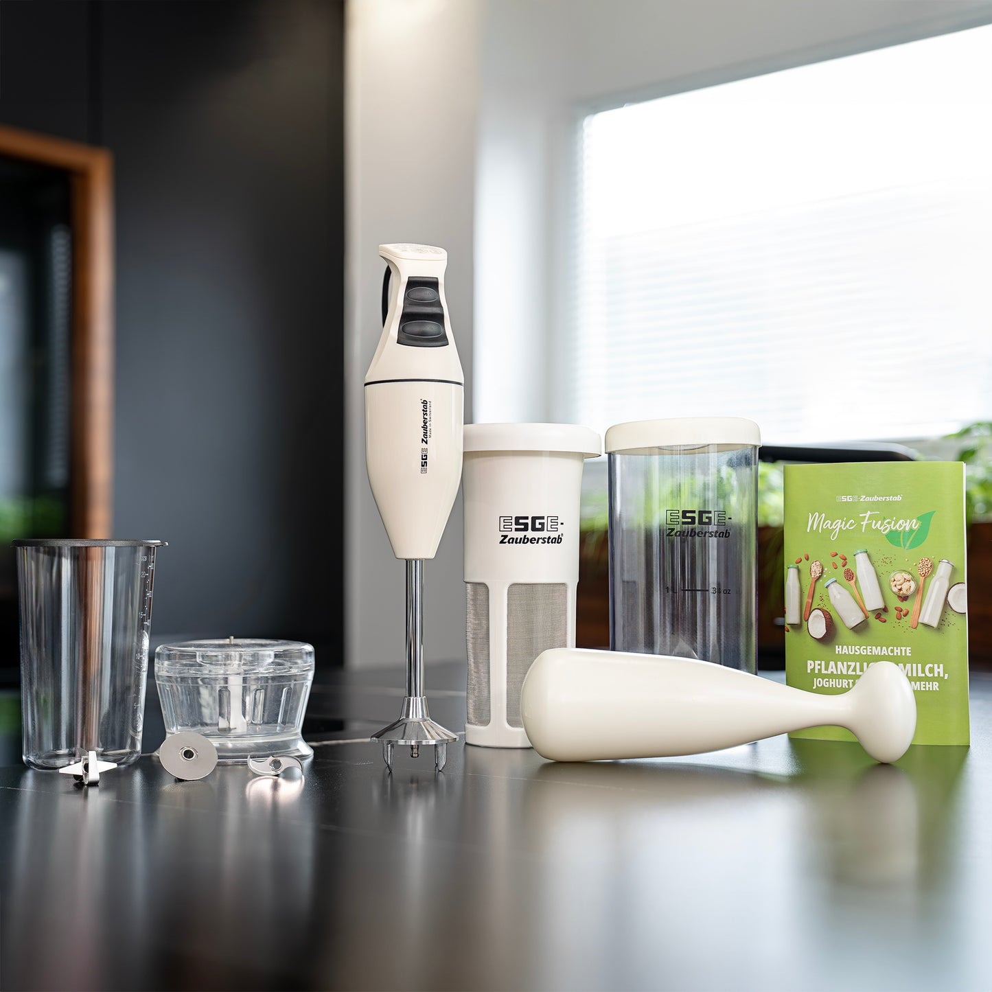 Der ESGE-Zauberstab E 120 Select + Magic Fusion Creme Stabmixer mit Aufsätzen und Behältern steht auf einer Küchentheke neben einer grünen Magic Fusion Creme-Box, während das Sonnenlicht durch das Fenster fällt.
