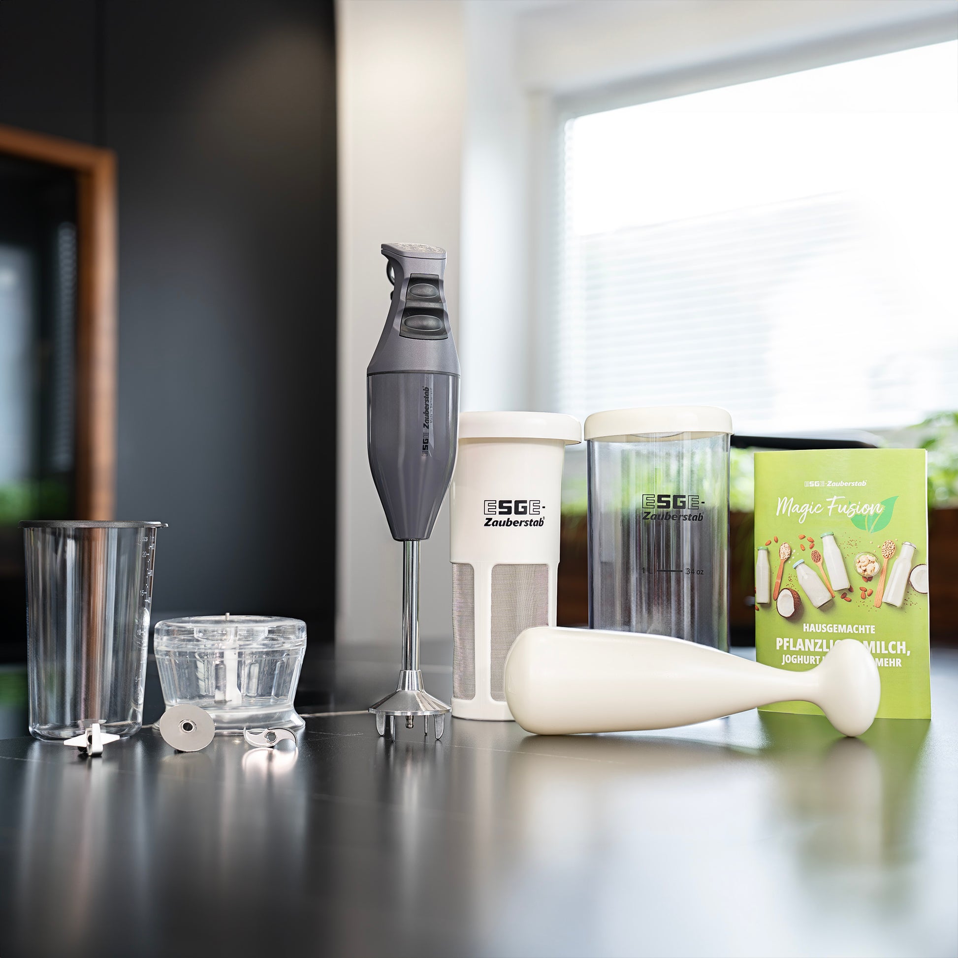 Auf einer Theke stehen der ESGE-Zauberstab E 120 Select von Unold mit Mischgläsern, Messbechern, einem weißen Stampfer und der Magic Fusion Anthrazit-Box mit pflanzlichen Milchgetränken.