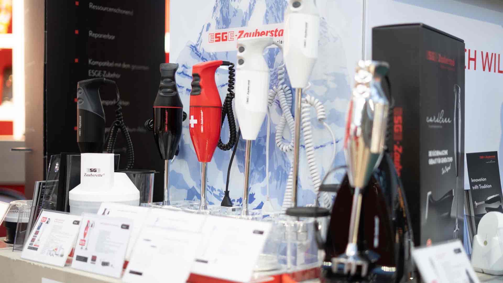 Eine Ausstellung verschiedener Stabmixer in unterschiedlichen Farben, darunter Schwarz, Rot und Weiß, angeordnet auf einer Theke mit Produktkartons und Informationskarten vor einem Hintergrund mit einem Bild eines schneebedeckten Berges.