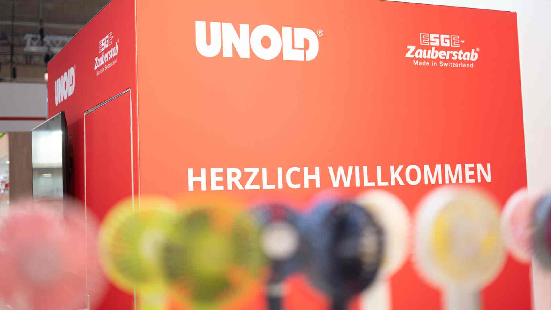 Ein roter Schaukasten mit den Aufschriften UNOLD und HERZLICH WILLKOMMEN sowie ESGE Zauberstab Made in Switzerland, im Vordergrund sind verschwommene bunte Gegenstände zu sehen.