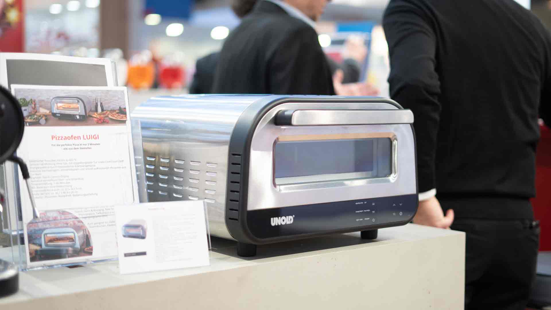 Bei einer Veranstaltung wird auf einer Theke ein kompakter Pizzaofen von UNOX ausgestellt. In der Nähe liegen Informationsbroschüren, im Hintergrund stehen zwei Personen in Geschäftskleidung.