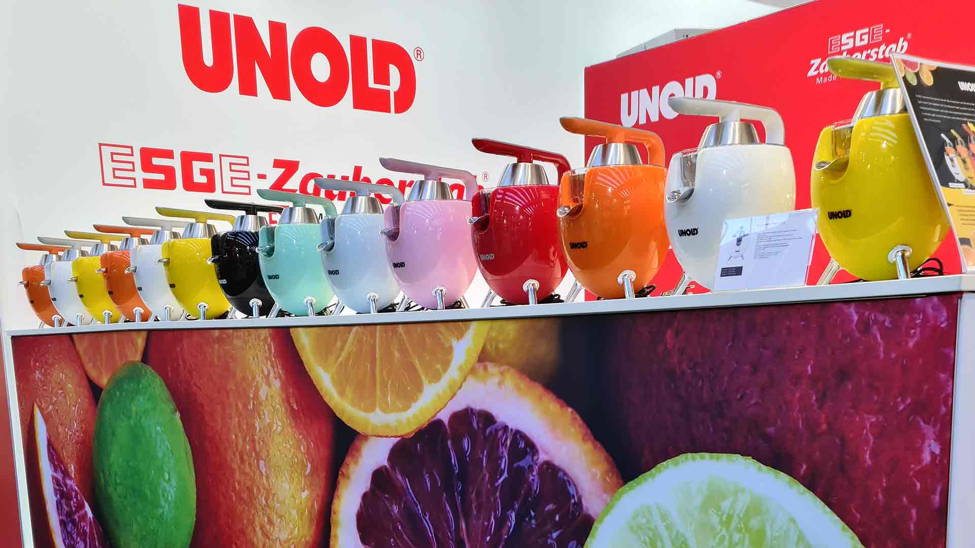 Auf einer Theke stehen mehrere bunte Mixer, jeder in einer anderen leuchtenden Farbe. Im Hintergrund sind ein großes UNOLD-Schild und Obstbilder zu sehen.