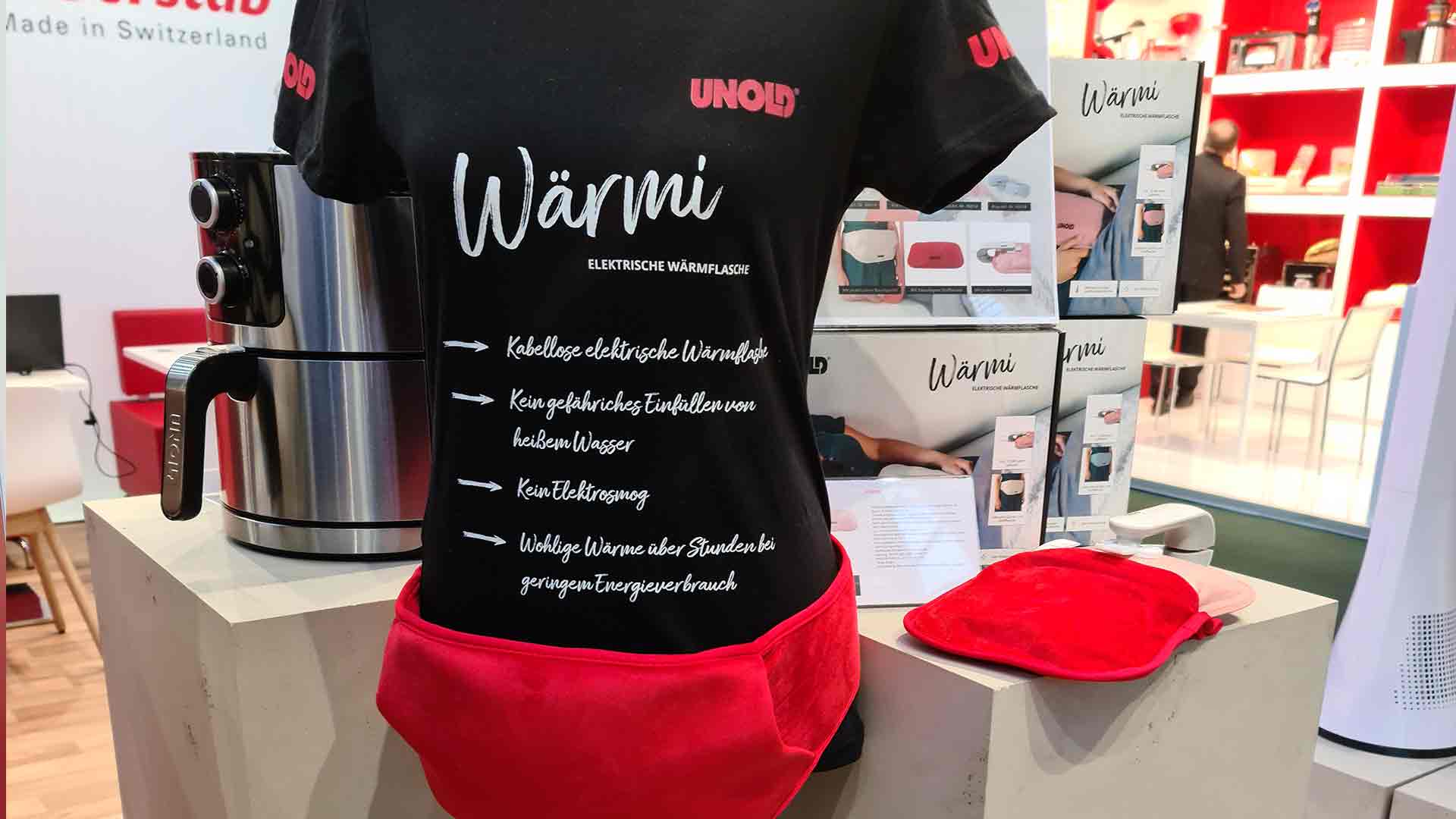 Eine Schaufensterpuppe trägt ein schwarzes Wärmi-T-Shirt und eine rote Gürteltasche an einem Messestand. Daneben stehen ein silbernes Haushaltsgerät und Heizkissen, im Hintergrund Produktregale und Werbedisplays.