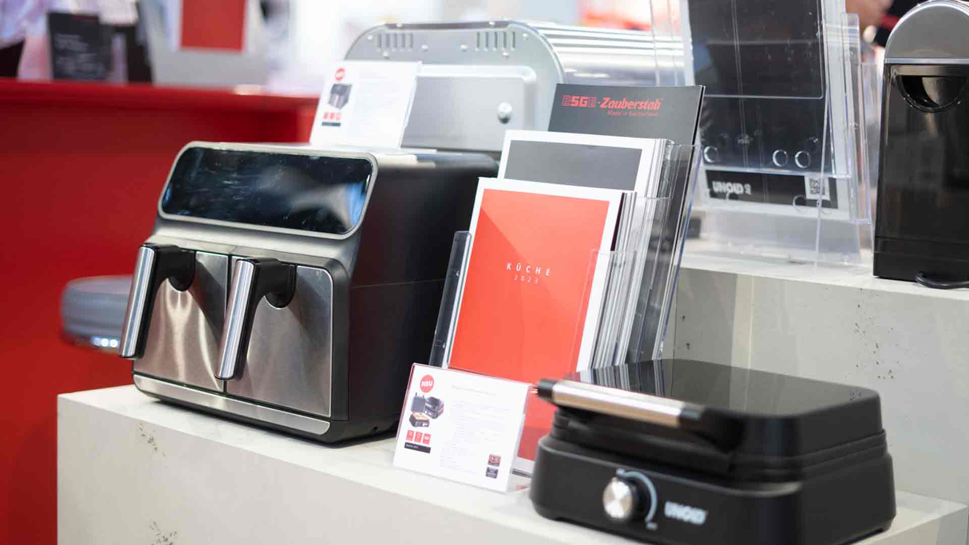Eine Ausstellungstheke mit einer modernen Heißluftfritteuse aus Edelstahl, Broschüren in Haltern und einem schwarzen Elektrogrill, alles vor einem rot-weißen Hintergrund.