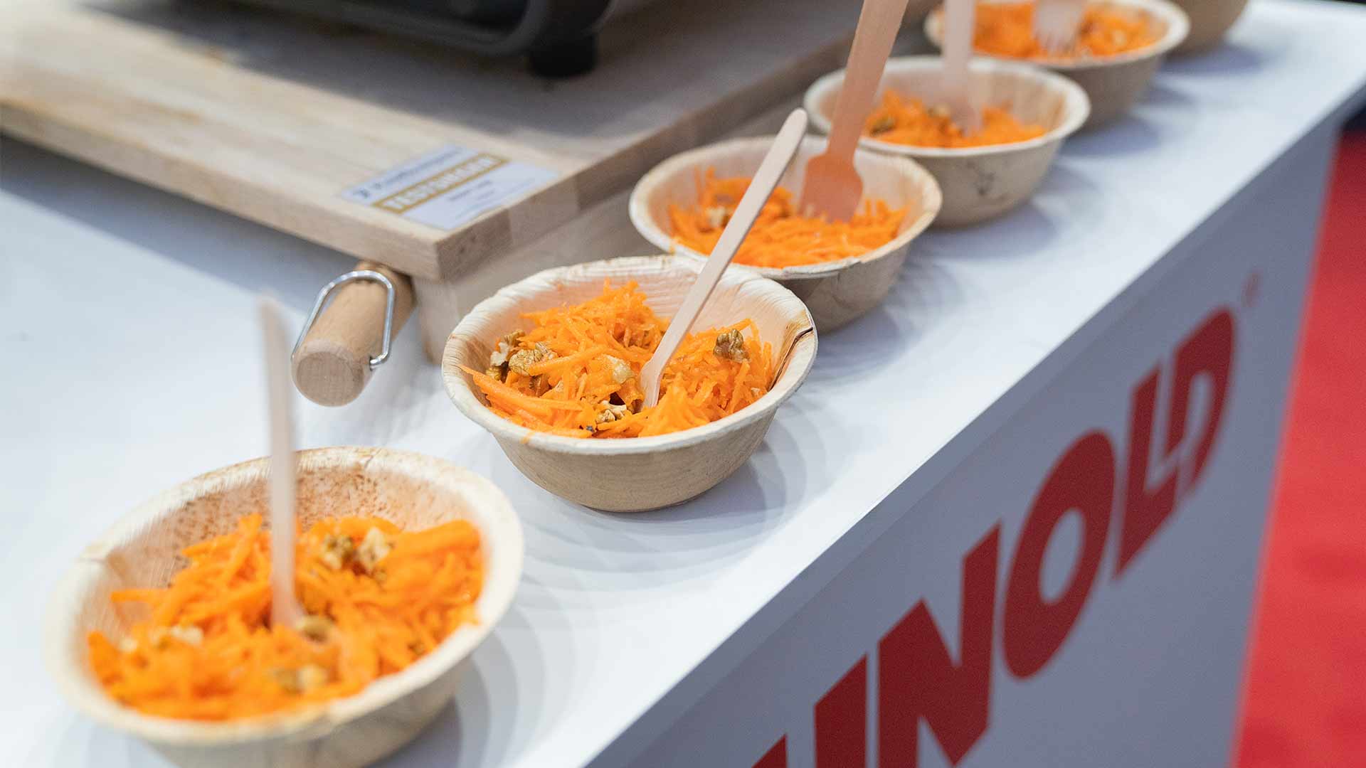 Fünf kleine Schüsseln mit geriebenen Karotten und Walnüssen stehen in einer Reihe auf einer Theke, jeweils mit einer Holzgabel. Im Hintergrund ist ein Schneidebrett zu sehen.
