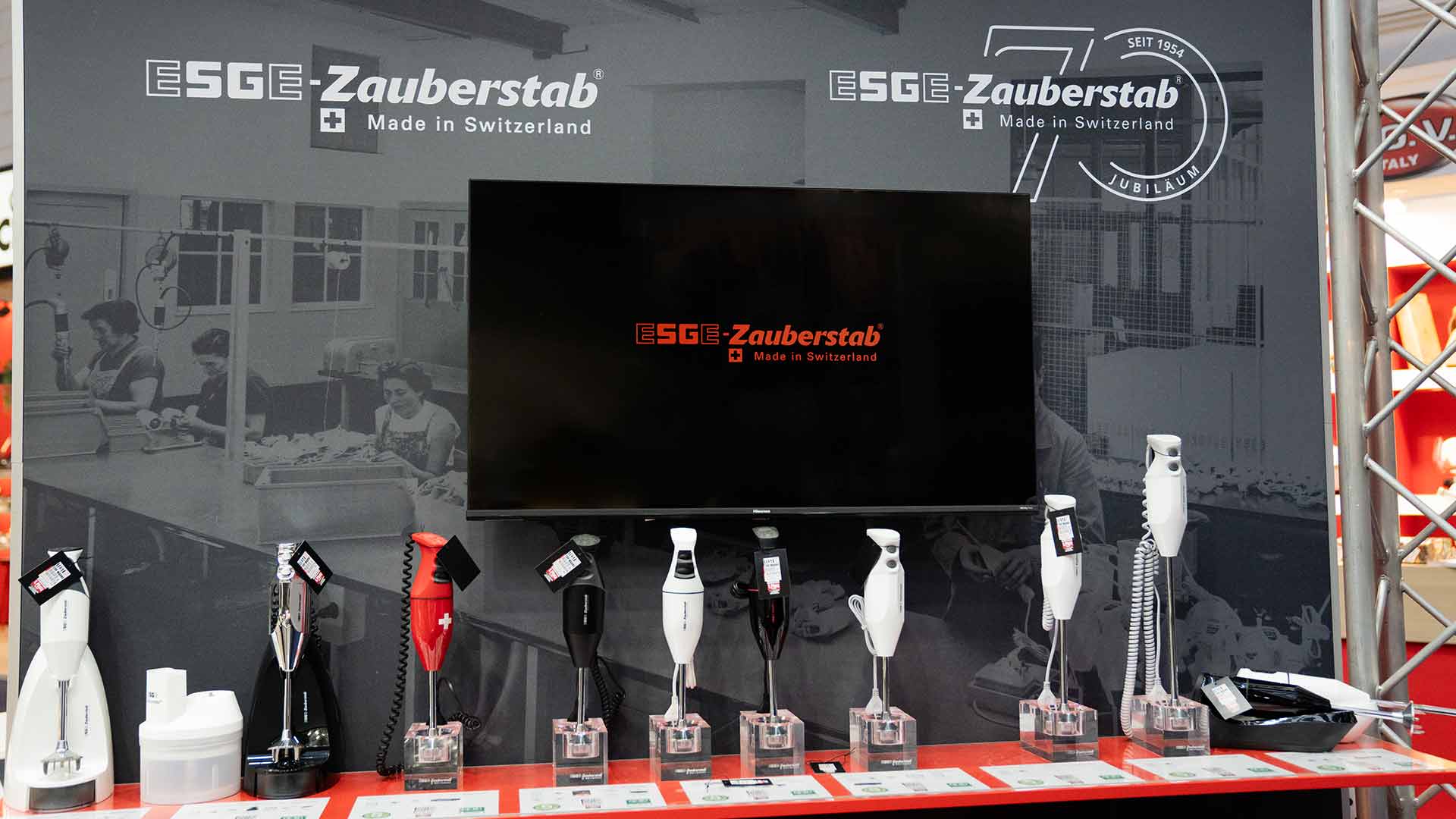 Auf einem Messestand sind acht Stabmixer und Zubehör vor einem großen Bildschirm und einer ESGE-Zauberstab-Markenbeschilderung ausgestellt.