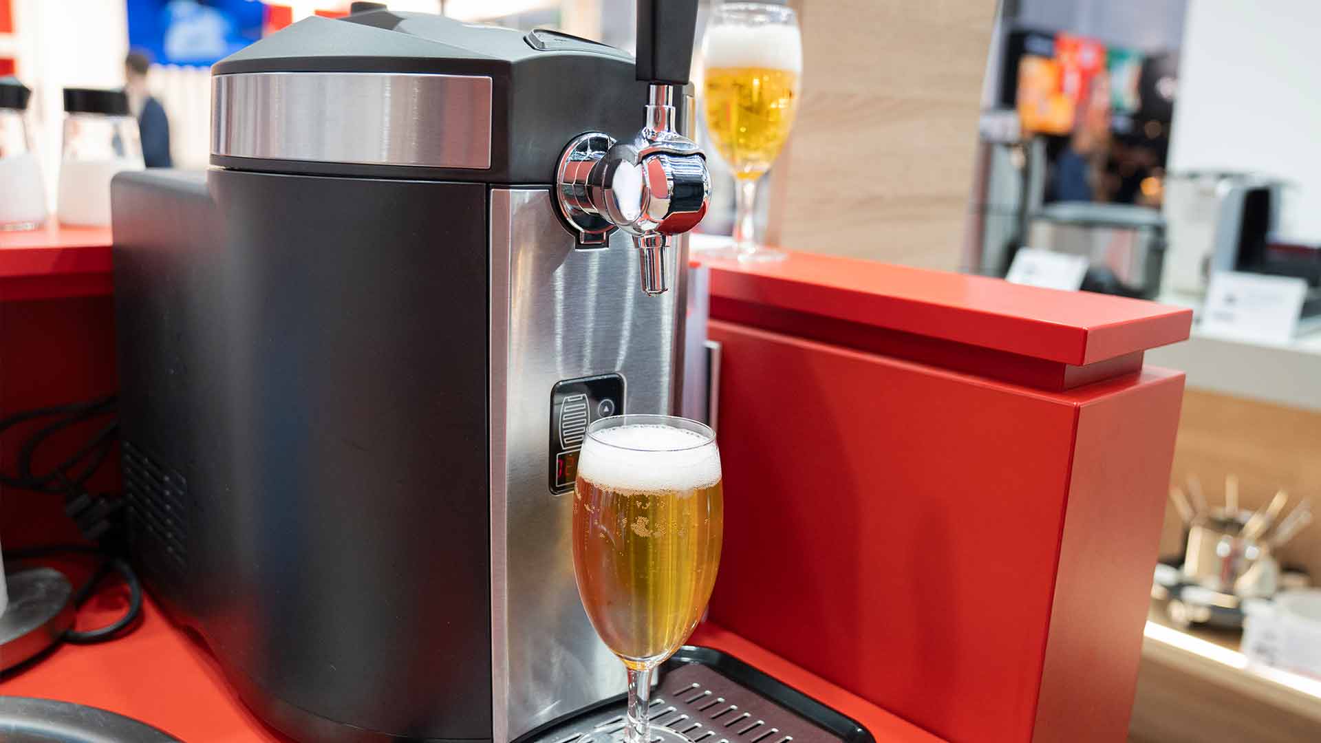 Nahaufnahme einer Bierzapfanlage, die Bier in ein Glas gießt, das auf einer Abtropfschale auf einer roten Theke steht. Das Bier hat Schaum. Im Hintergrund sind unscharfe Objekte und Personen zu sehen.