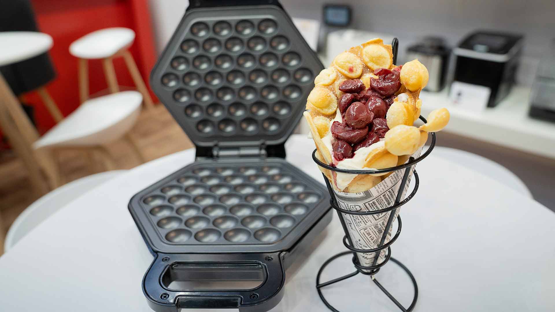 Auf einem Tisch steht ein Bubble-Waffel-Eisen, geöffnet neben einer Bubble-Waffel-Tüte, gefüllt mit Schlagsahne und Kirschen, die in einem mit Zeitungspapier ausgelegten Metallständer präsentiert wird.
