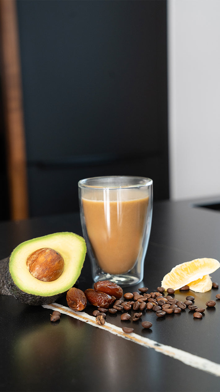 Auf einer schwarzen Fläche steht ein Glas Kaffee, umgeben von einer halben Avocado, einer Zitronenscheibe, Datteln und verstreuten Kaffeebohnen, vor einem dunklen Hintergrund.