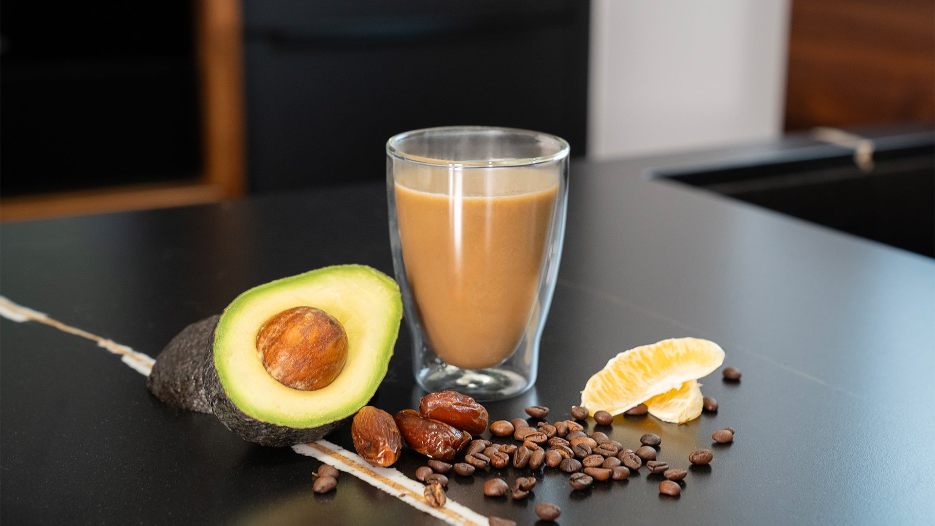 Glas mit Kaffee auf einer Küchenzeile, daneben liegen zwei Stücke Orange, 3 Datteln, eine halbe Avocado mit Kern und eine Hand voll Kaffeebohnen.